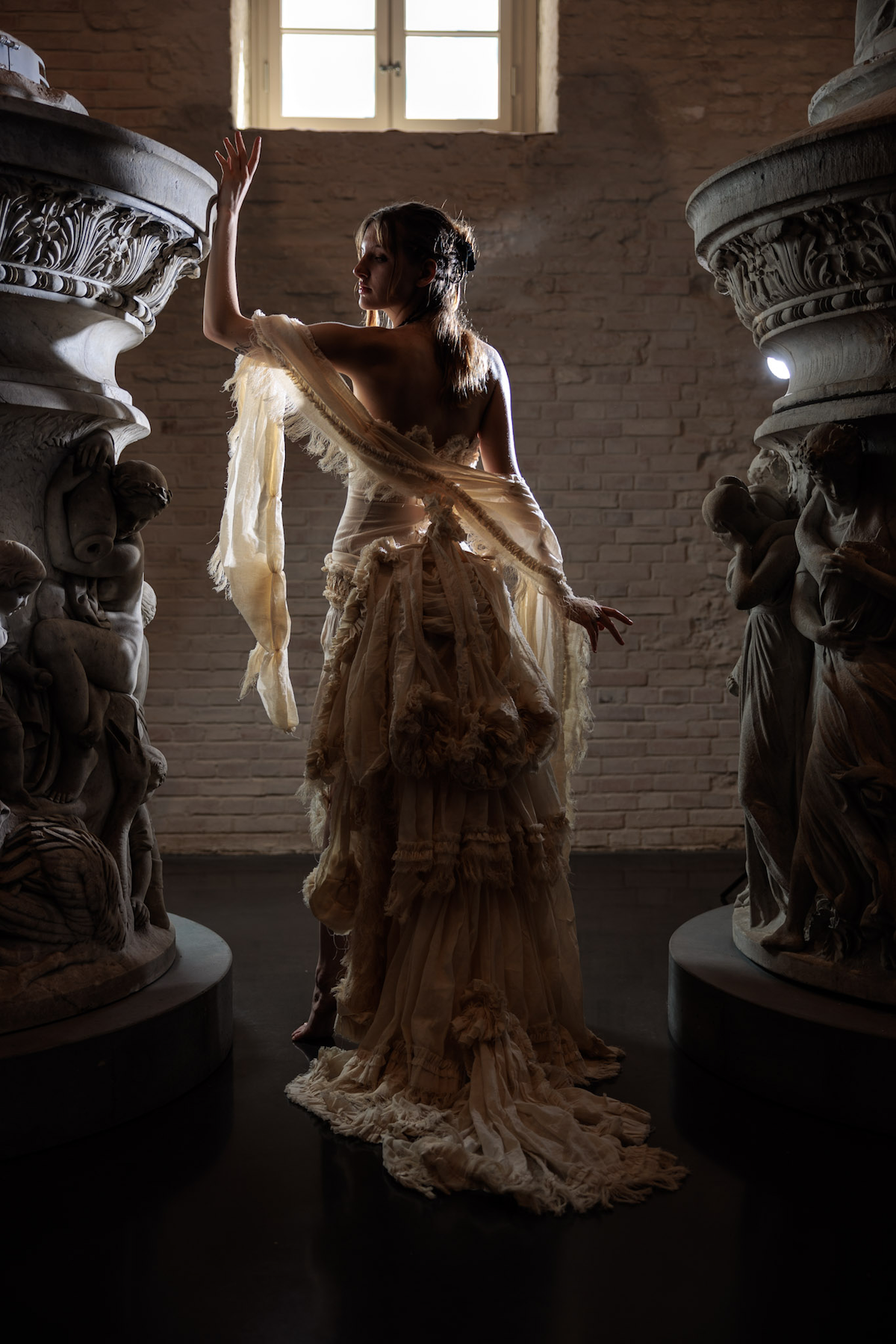 Erinnerungsstück-Janiejean-Couture-Frau in einem langen, zerzausten, alten Kleid steht zwischen zwei antiken Skulpturen im Licht eines Fensters.
