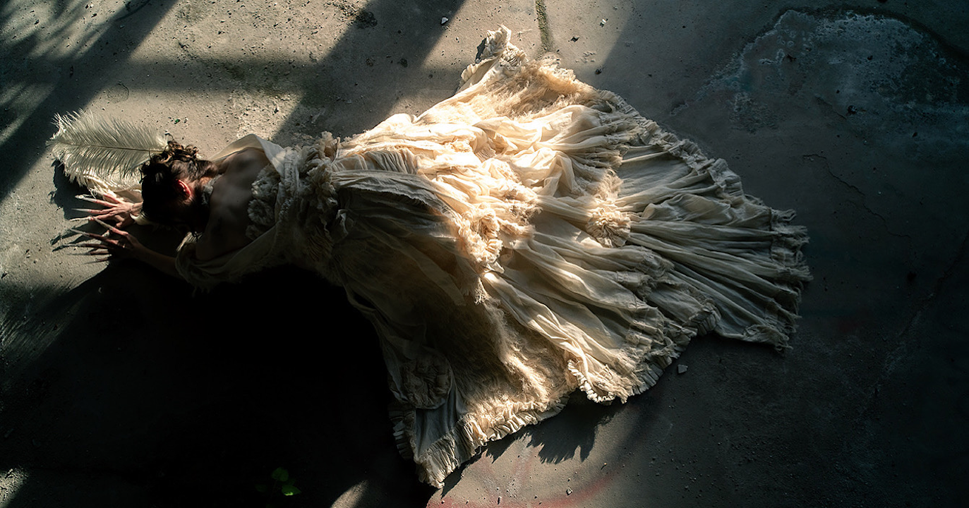 Janiejean-Haute-Couture,Langes schleppen Kleid-couture-Vogelperspektive-langgestreckt-Feder- fFrau in einem antiken, zerknitterten Kleid mit Federschmuck liegt auf dem Boden, in schattigem Licht.