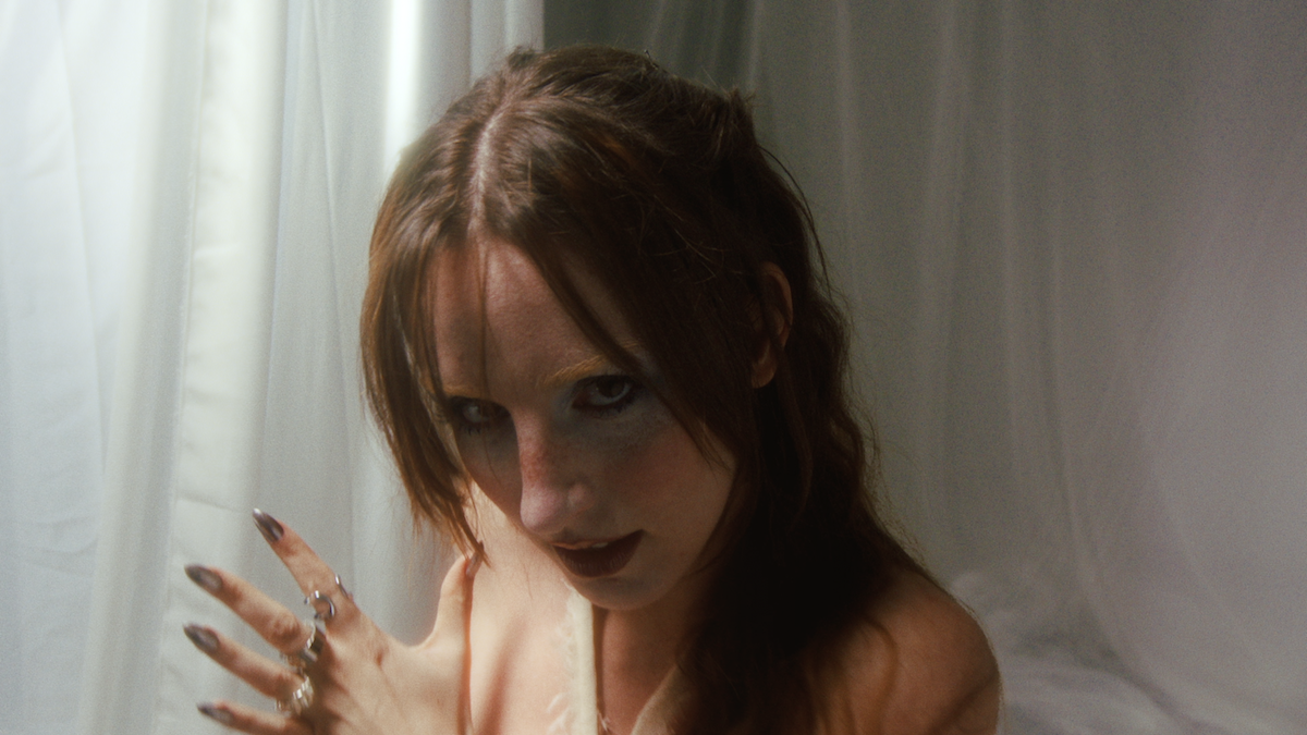 Nahaufnahme einer Frau mit roten Haaren und dunklem Make-up, die vor weißen Vorhängen steht.