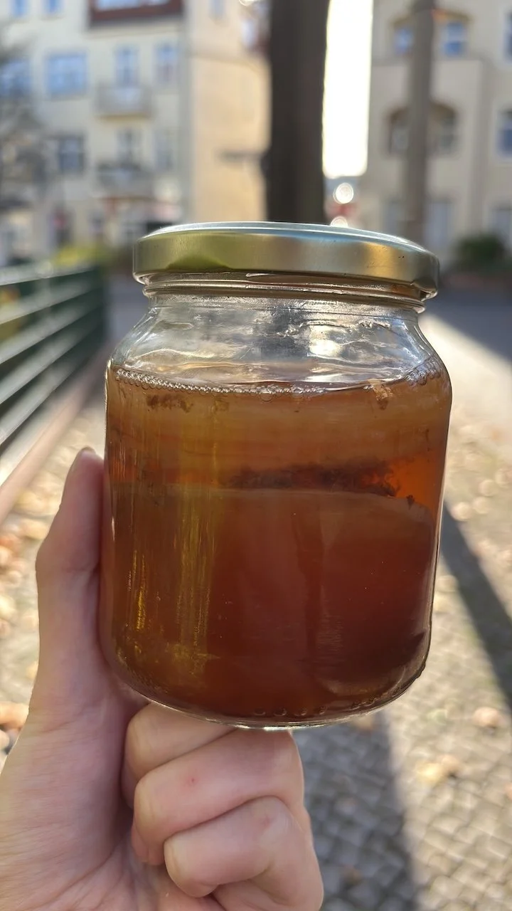 Janiejean-Scoby-Textile-Surfaces, Ein Glas mit dunkler brauner Substanz, vermutlich Tee oder Fruchtmarmelade, in der Hand vor einem Stadtbild mit Mehrfamilienhäusern und einem Gehweg im Freien.