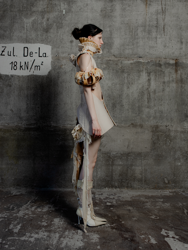 Eine Frau trägt ein avantgardistisches, modisches Outfit mit großen Rüschen und einer neutralen Farbpalette in einem grauen, industriellen Raum.