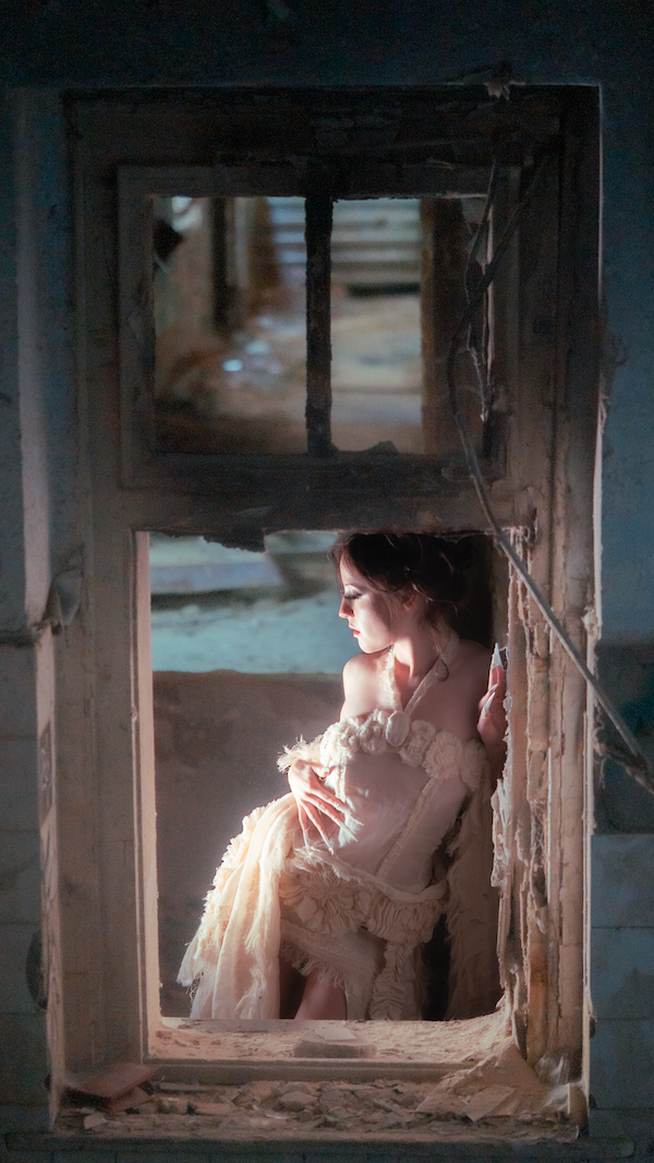 Erinnerungsstück-Janiejean-Couture-Junge Frau sitzt hinter einem alten, zerbrochenen Fensterrahmen in einem Raum, getragen in einem weißen, flauschigen Kleid, mit Sonnenlicht, das sie beleuchtet.