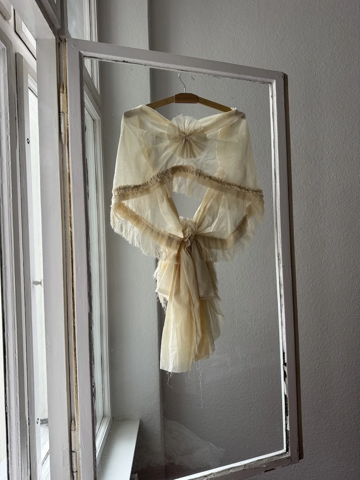 Erinnerungsstück-Janiejean-Couture-Ein Vintage-Spitzen-Umhang in Cremefarbe, der an einem Holzkleiderbügel im Rahmen eines großen Fensters hängt.