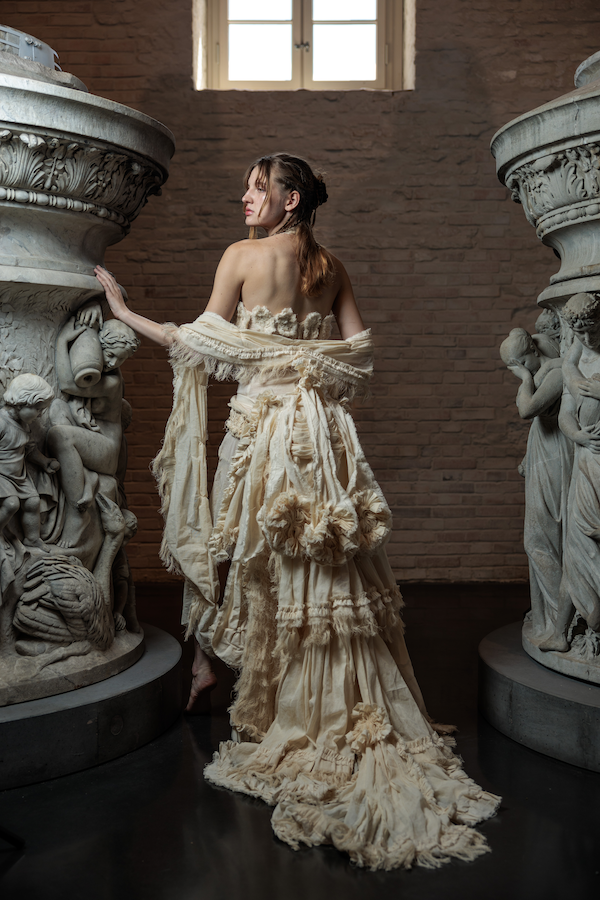 Erinnerungsstück-Janiejean-Couture-Junge Frau in einem eleganten, cremefarbenen Kleid zwischen zwei kunstvollen, antiken Säulen im Museum, mit einem Fenster im Hintergrund.