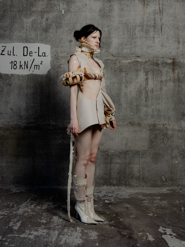 Eine Frau steht in einem minimalistischen, grauen Raum. Sie trägt eine avantgardistische, beige und kragenartige Kleidung mit hohen Stiefeln.