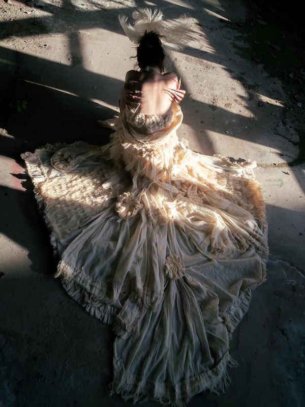 Erinnerungsstück-Janiejean-Couture-Frau in einem Vintage- Kleid mit Federn auf dem Kopf sitzt auf einer Steinboden im Schatten, Blick nach unten.