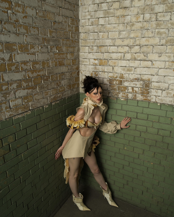 Eine Frau in einem ausgefallenen Outfit mit hohen Schuhen steht in einer Ecke mit braunen und grünen Ziegelwänden.