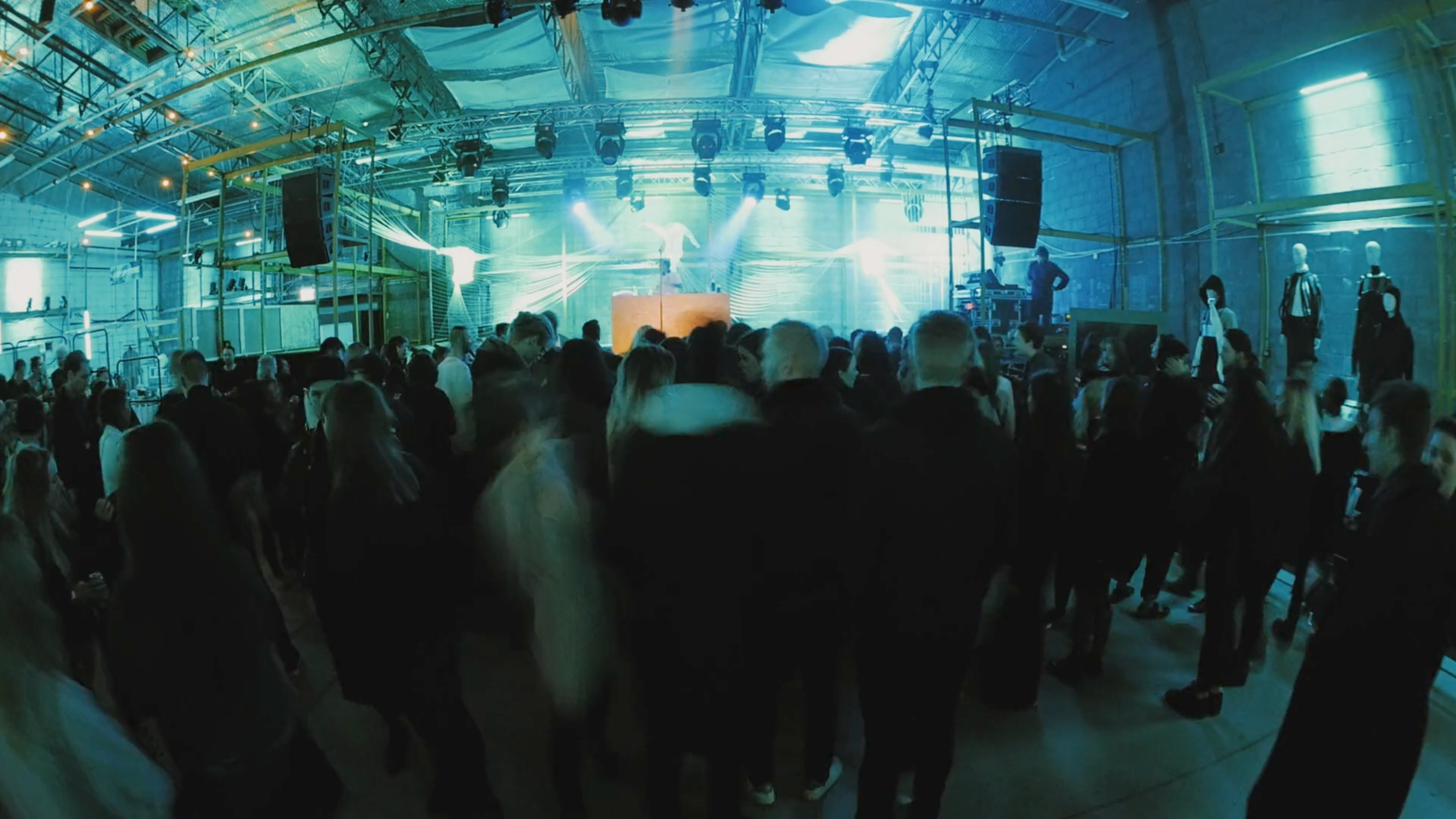 People dancing and socializing at a nightclub with a DJ performing on stage, blue lighting, mannequins on racks, and industrial decor.