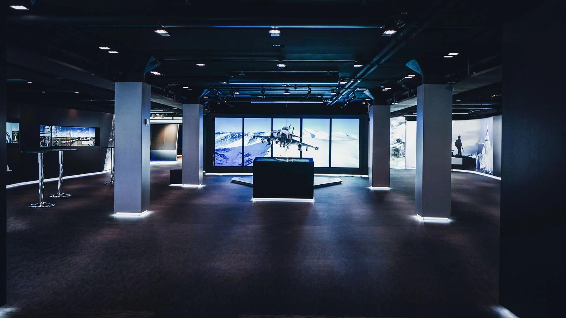Exhibition room with a central display of a fighter jet model in front of a large screen showing mountains and a fighter jet in flight.