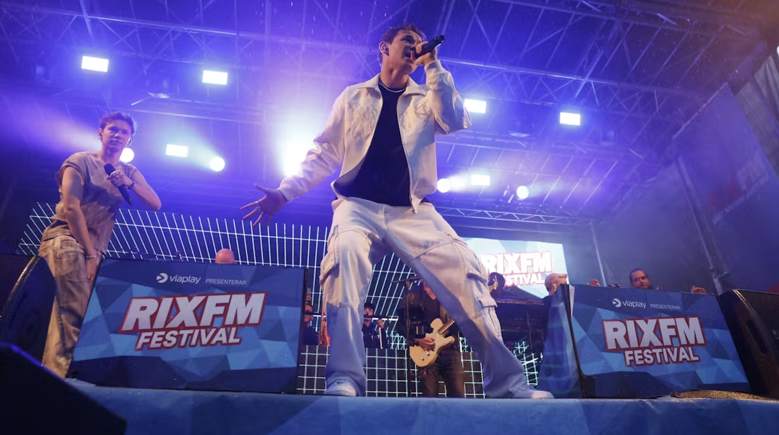 Musicians performing at the RIX FM Festival, with a singer in the foreground holding a microphone and a guitarist in the background. The stage is illuminated with purple and blue lights.