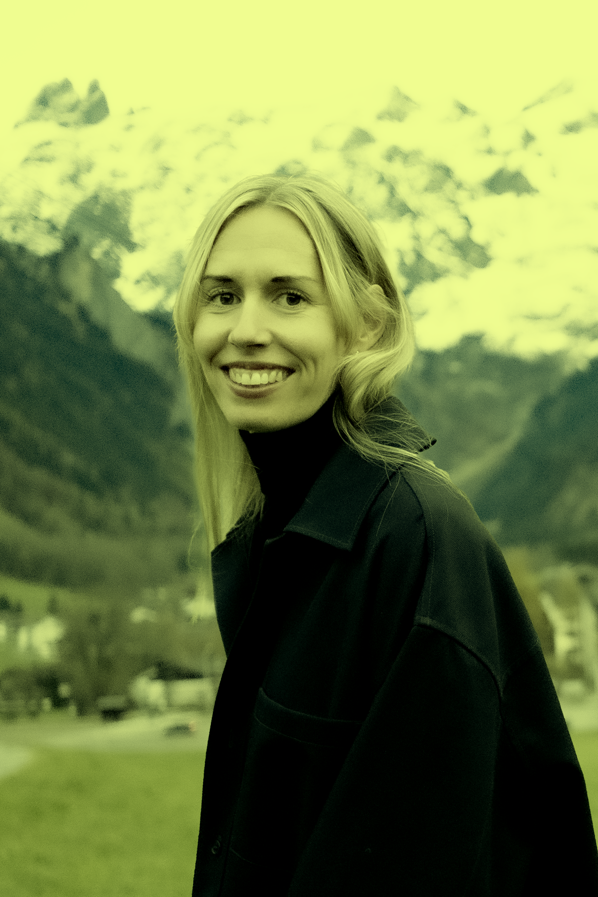 A smiling woman with blonde hair standing outdoors, with mountains and cloudy sky in the background.