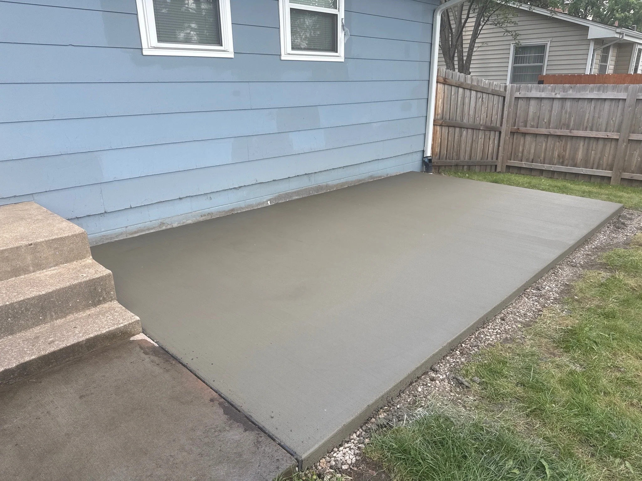 Freshly poured concrete slab in backyard next to a blue house with stairs and a wooden fence.