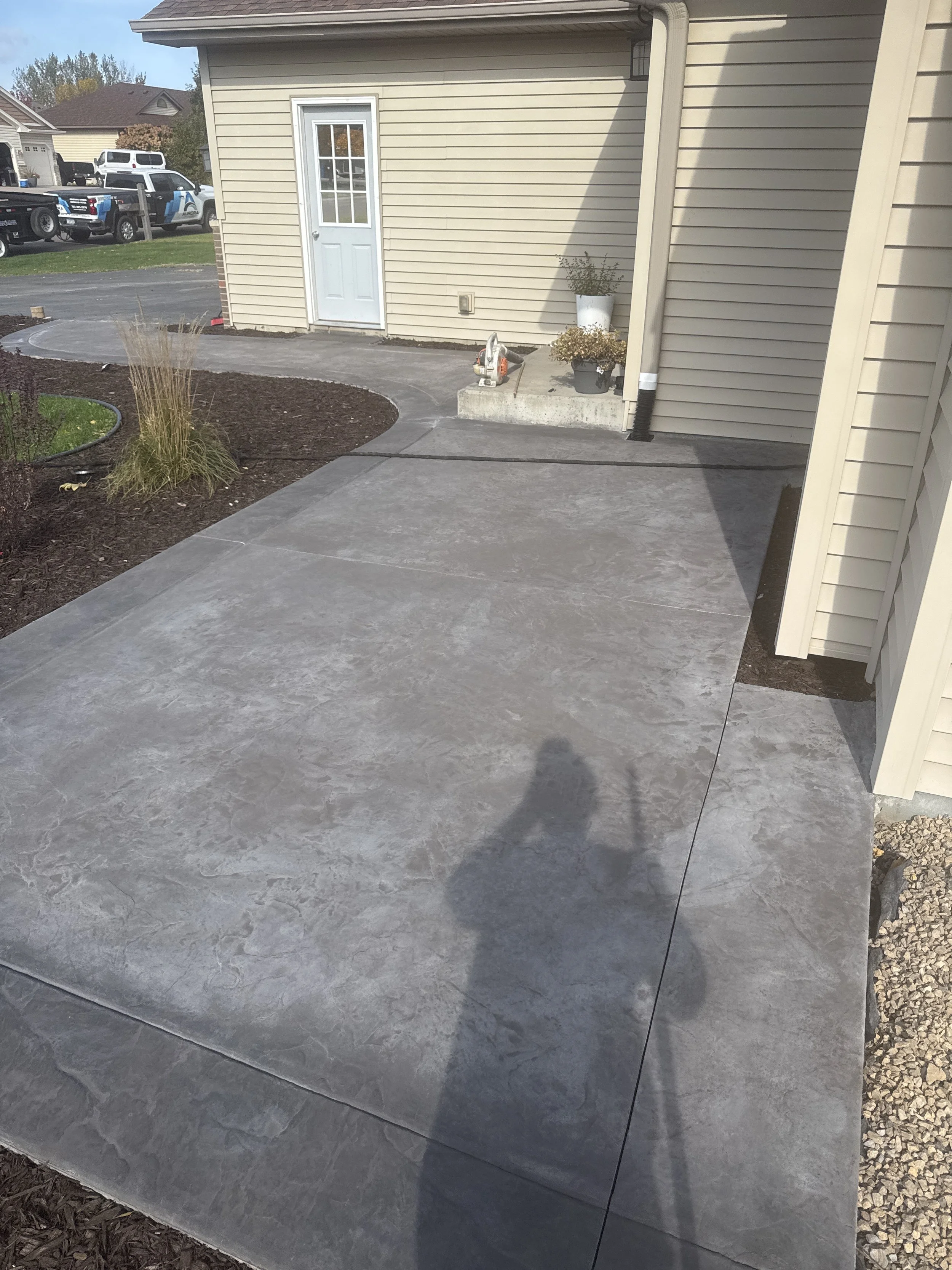 Newly poured concrete patio and sidewalk next to a house with beige siding, white trim, and a white door. Landscaping includes mulch and some plants, with a few vehicles parked nearby.