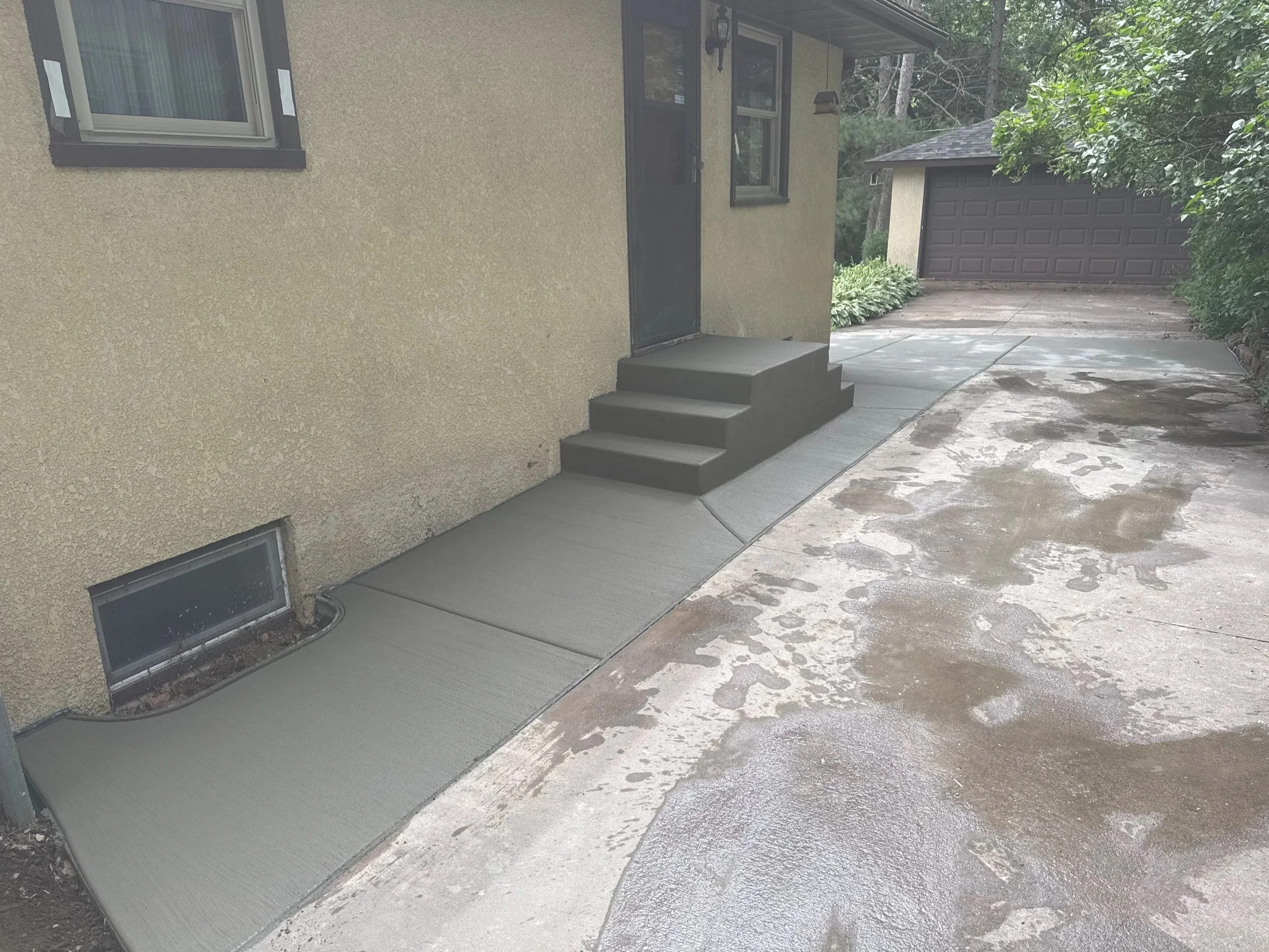 Concrete pathway leading to a front door with stairs, part of a house exterior with beige stucco wall, windows, and a garage in the background, surrounded by some greenery.