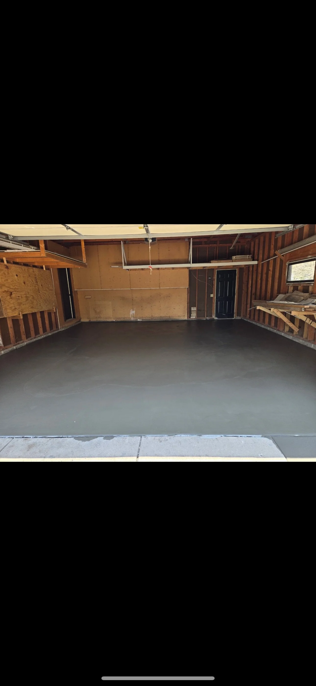 Empty garage with freshly poured concrete floor and wooden walls, black door, window, and some shelves on the right side.