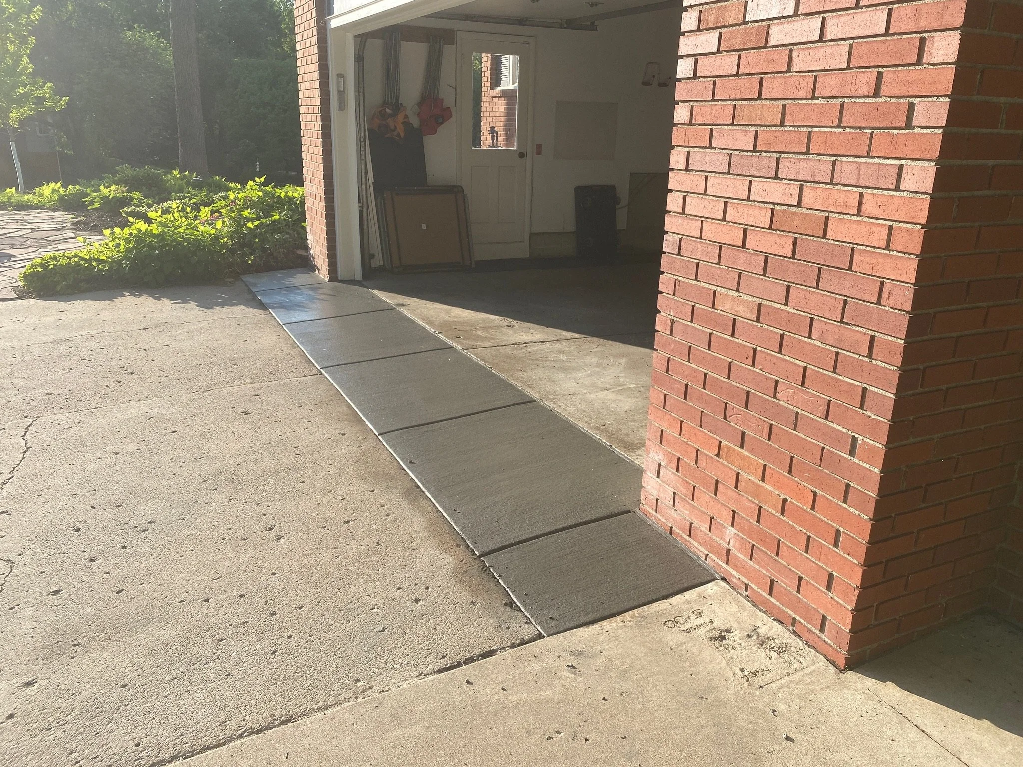 Concrete sidewalk leading to an open garage door on a sunny day. The garage has tools hanging on the wall and some items leaning against the wall inside.