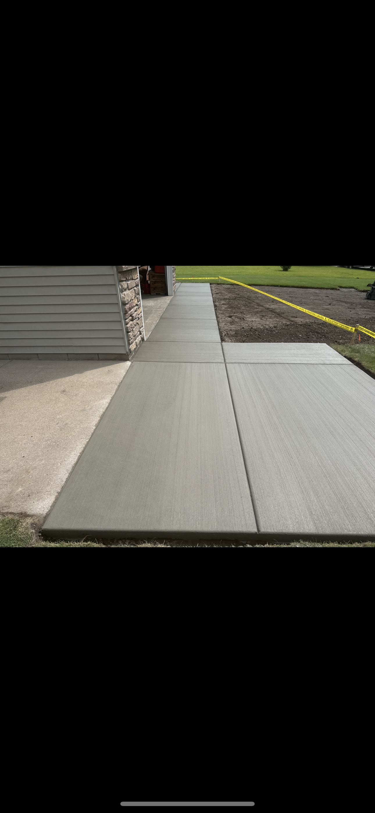 A newly poured concrete sidewalk adjacent to a house with vinyl siding and stone accents, with a grassy lawn and yellow caution tape in the background.