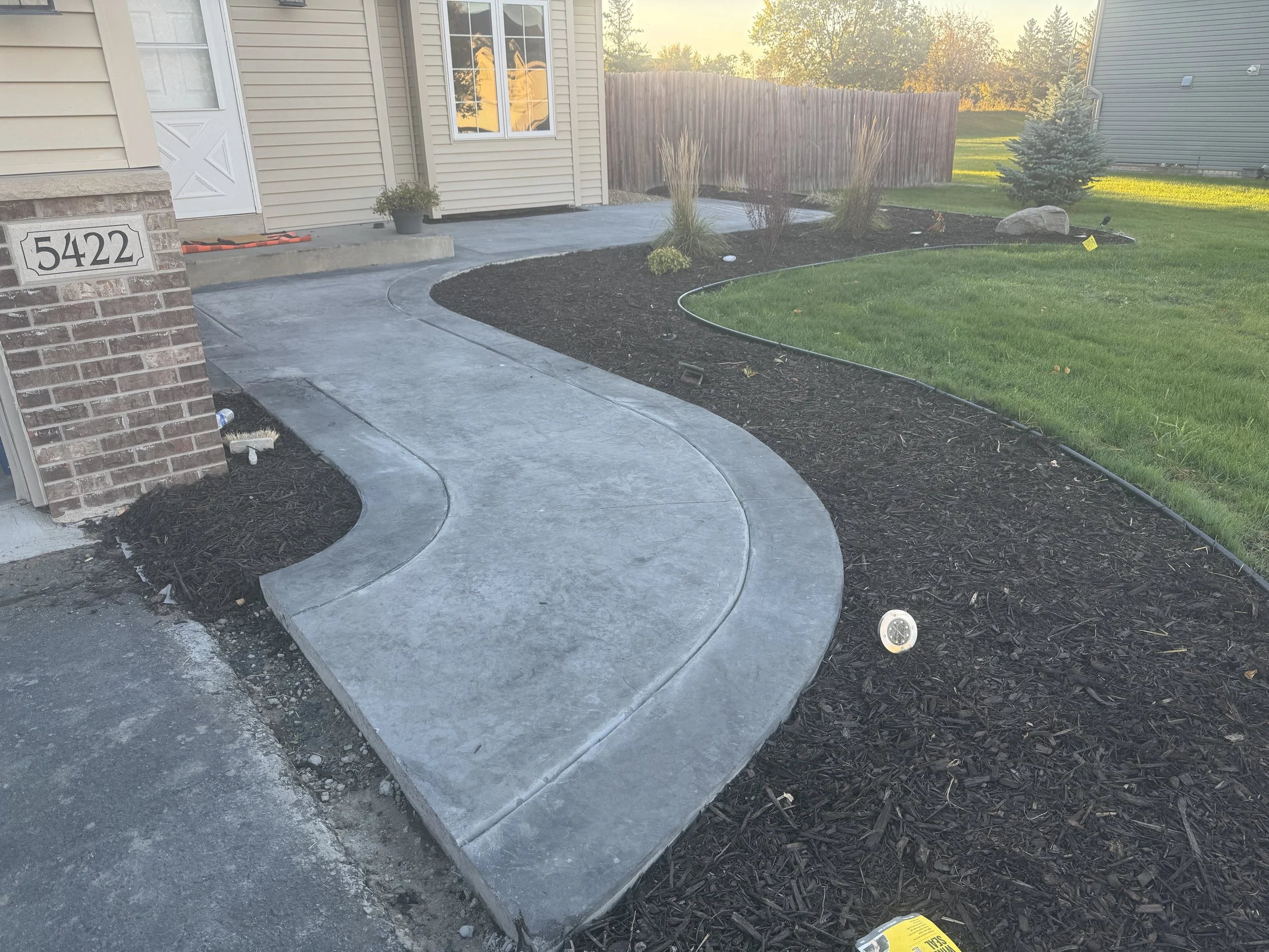 Newly poured concrete sidewalk and pathway in front of a house with a red brick front and beige siding, with a landscaped yard featuring plants, mulch, a house number plaque, and a grassy lawn illuminated by sunlight.