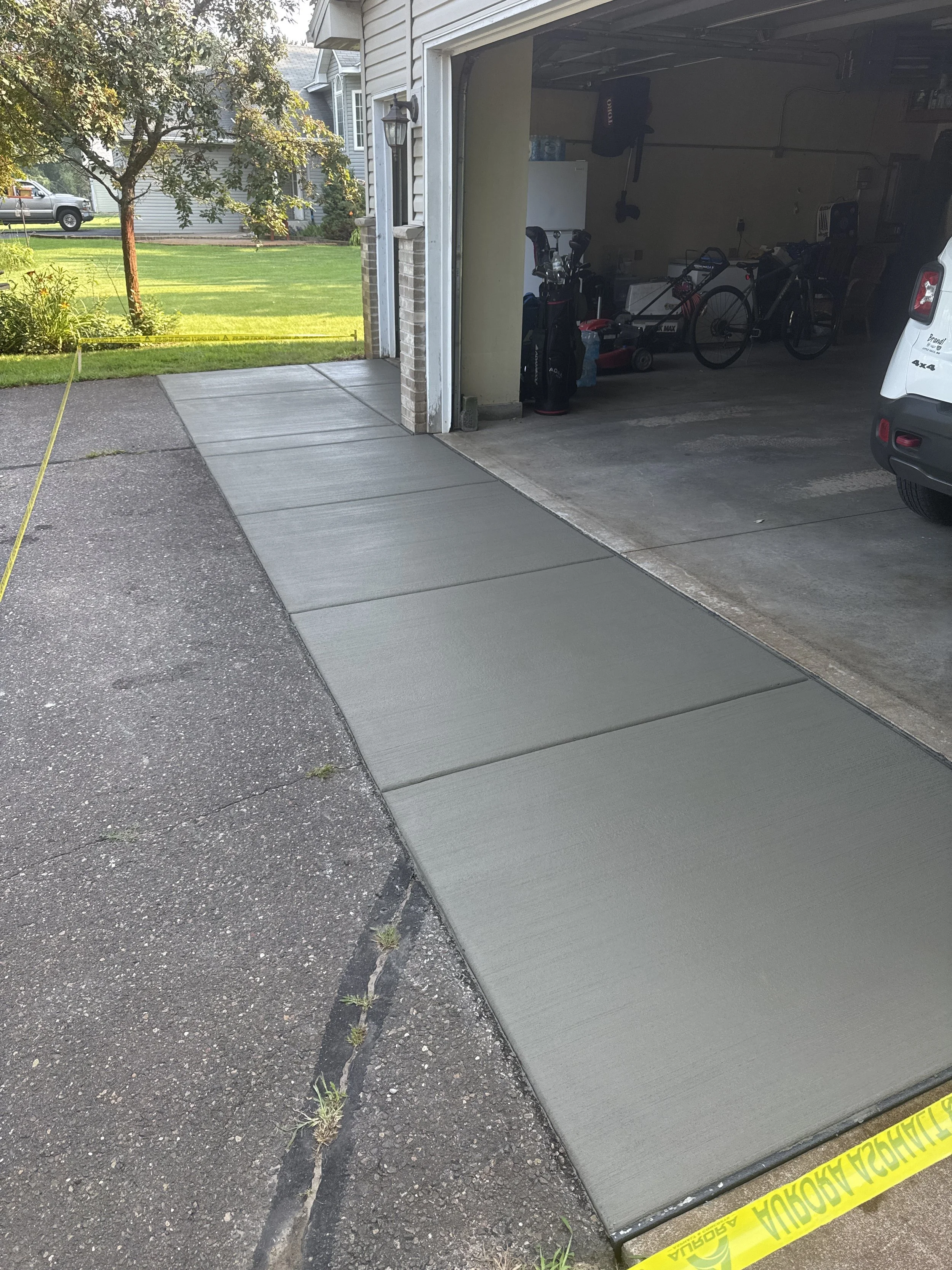Freshly poured concrete sidewalk in front of a garage, with yellow caution tape nearby, adjacent to a paved driveway, with a grassy yard and trees in the background.
