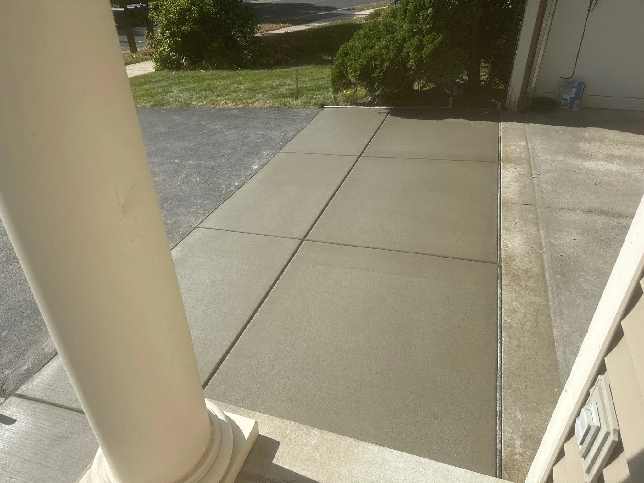 Concrete sidewalk and driveway leading to a house garage, with a white porch column in the foreground and bushes in the background.