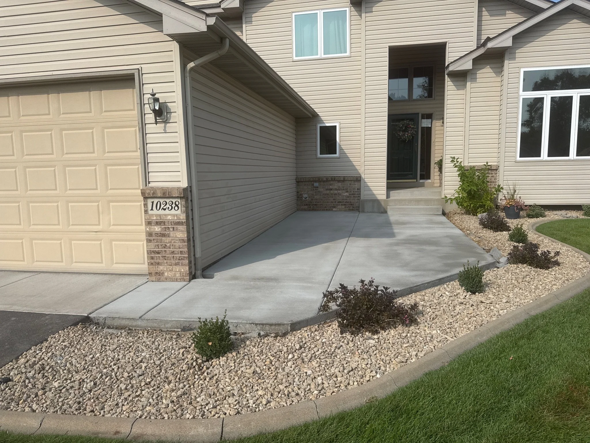 Concrete driveway and walkway leading to front door of beige siding house with brick accents and landscaped yard.