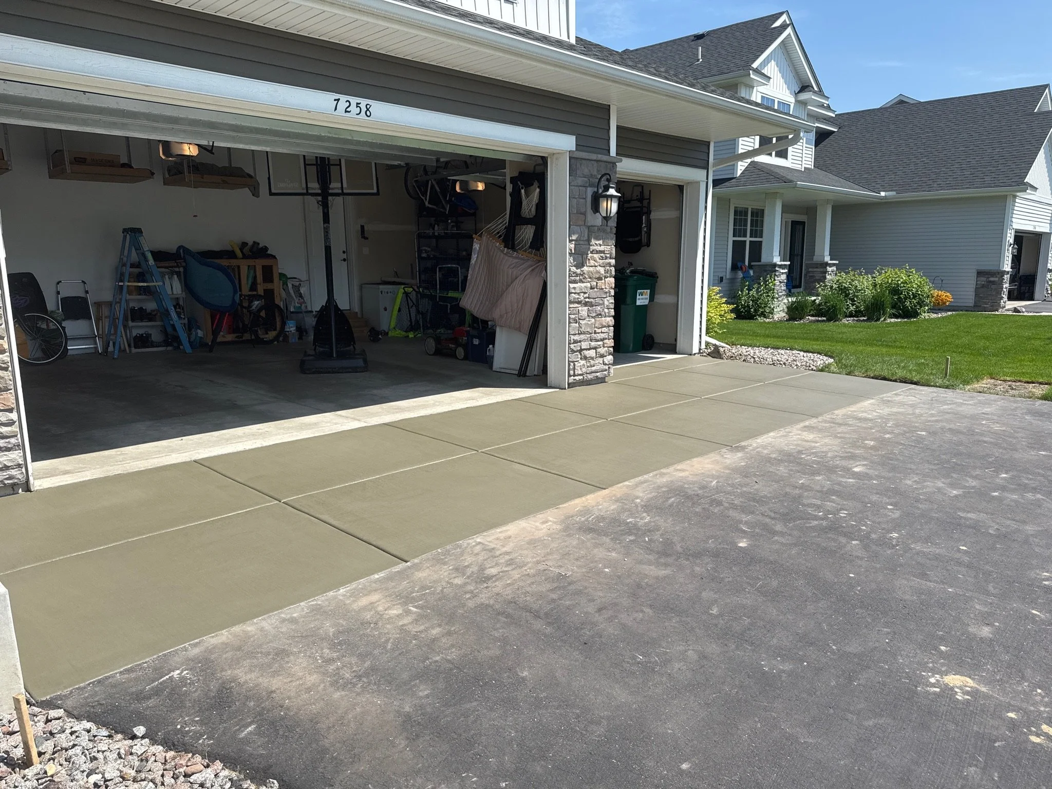 View of a house garage with open door, showing indoor storage items, and freshly laid concrete driveway outside on sunny day.