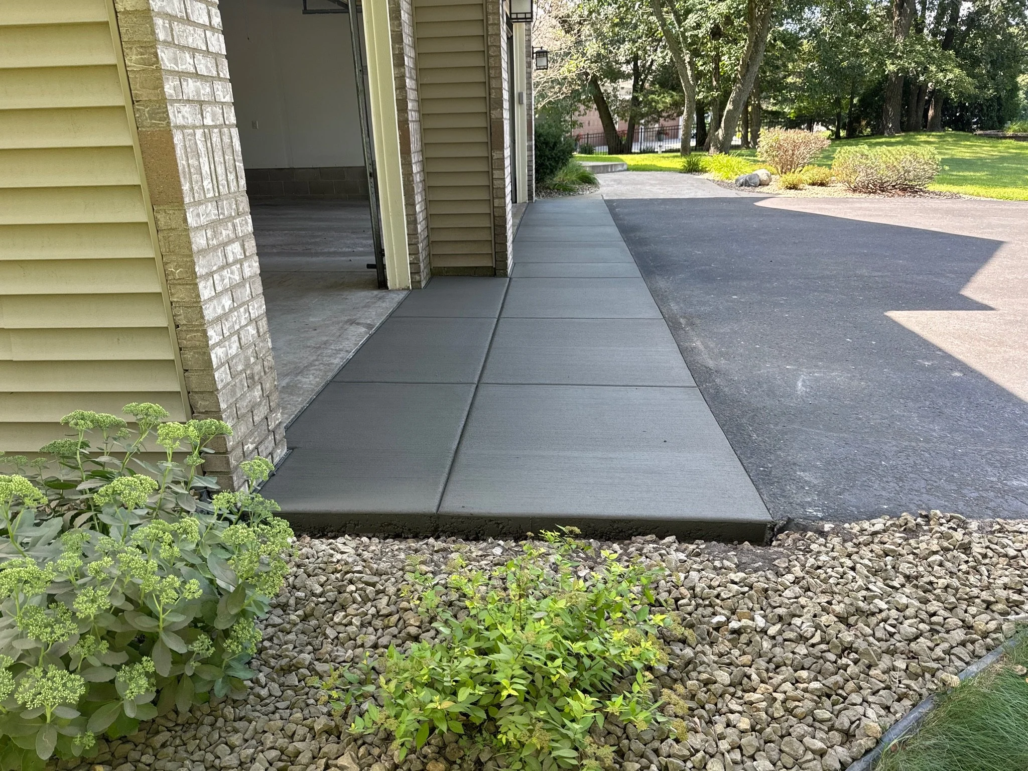 Concrete sidewalk next to a garage door, with gravel and plants in the foreground, and trees and lawn in the background.