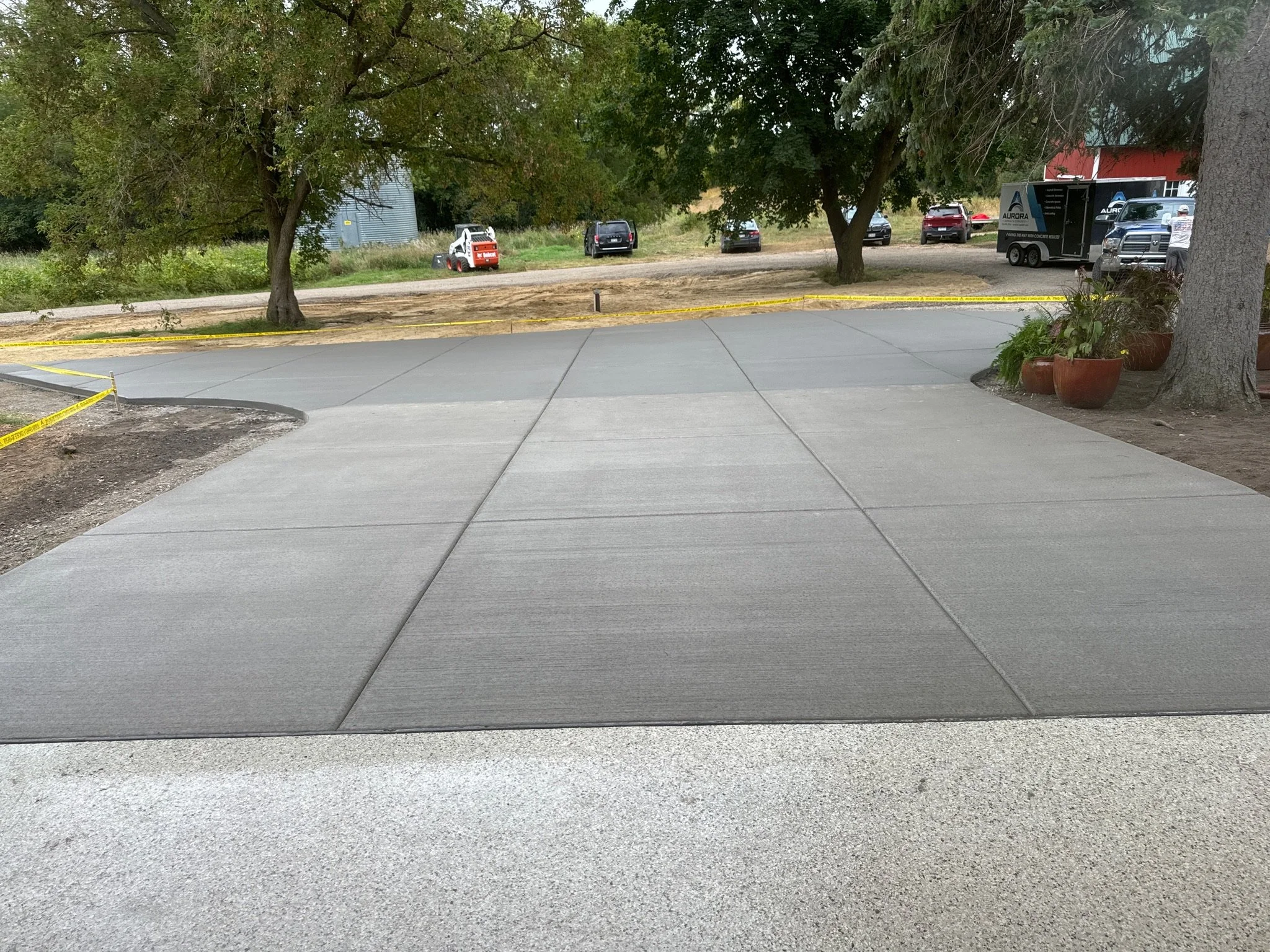 Freshly poured concrete sidewalk with visible expansion joints, surrounded by trees, plants, and parked vehicles, including a trailer and a small utility vehicle.