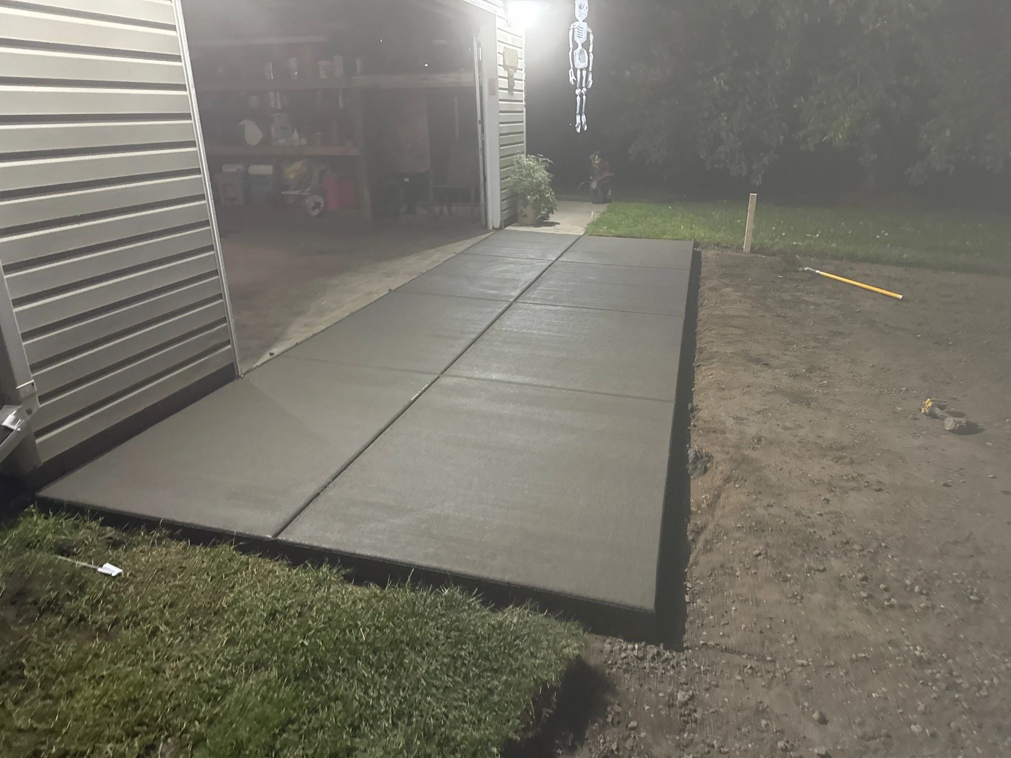 newly poured concrete sidewalk leading to garage with open metal door, interior storage visible, and a skeleton decoration hanging from the ceiling outside at night.