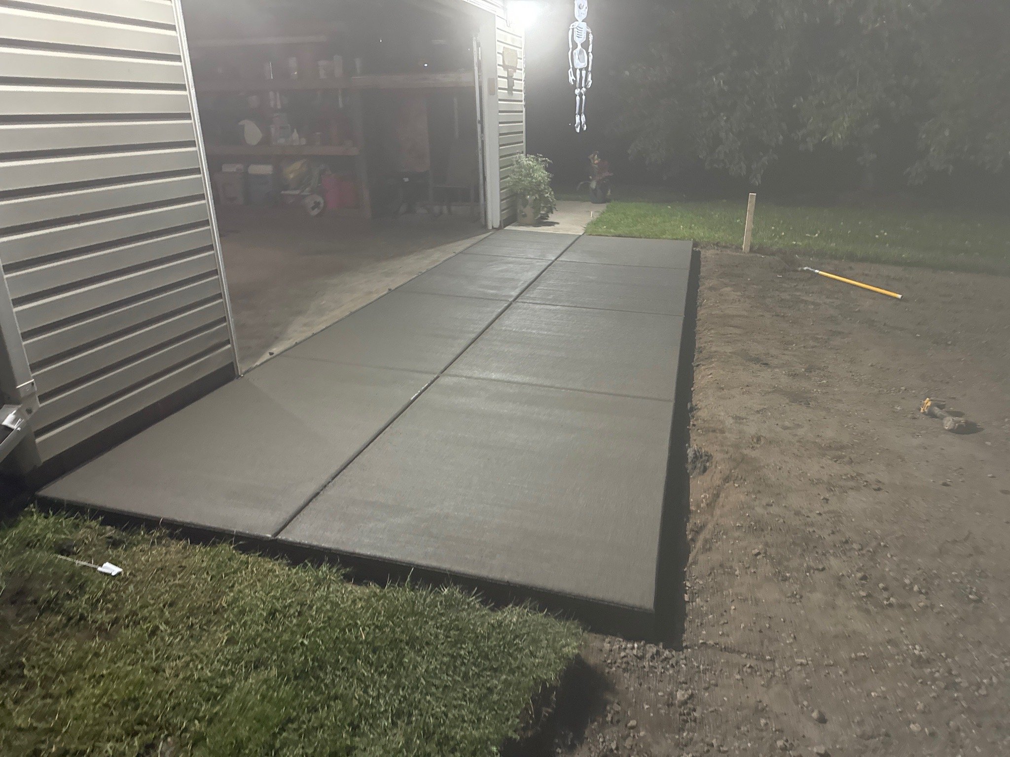 newly poured concrete sidewalk leading to garage with open metal door, interior storage visible, and a skeleton decoration hanging from the ceiling outside at night.