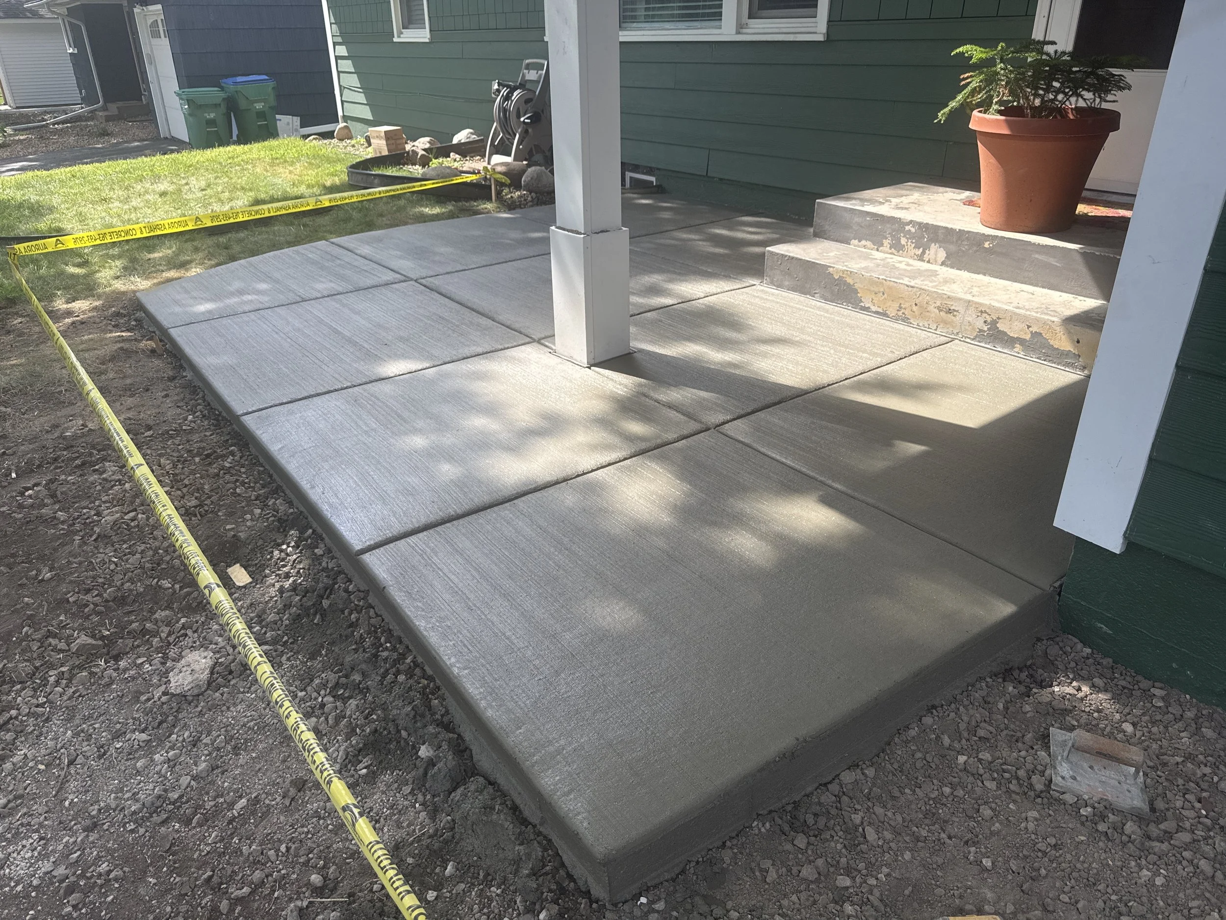 Newly poured concrete porch with large square slabs in front of a house with green siding, a small set of steps leading up to a door, a potted plant, and caution tape nearby.