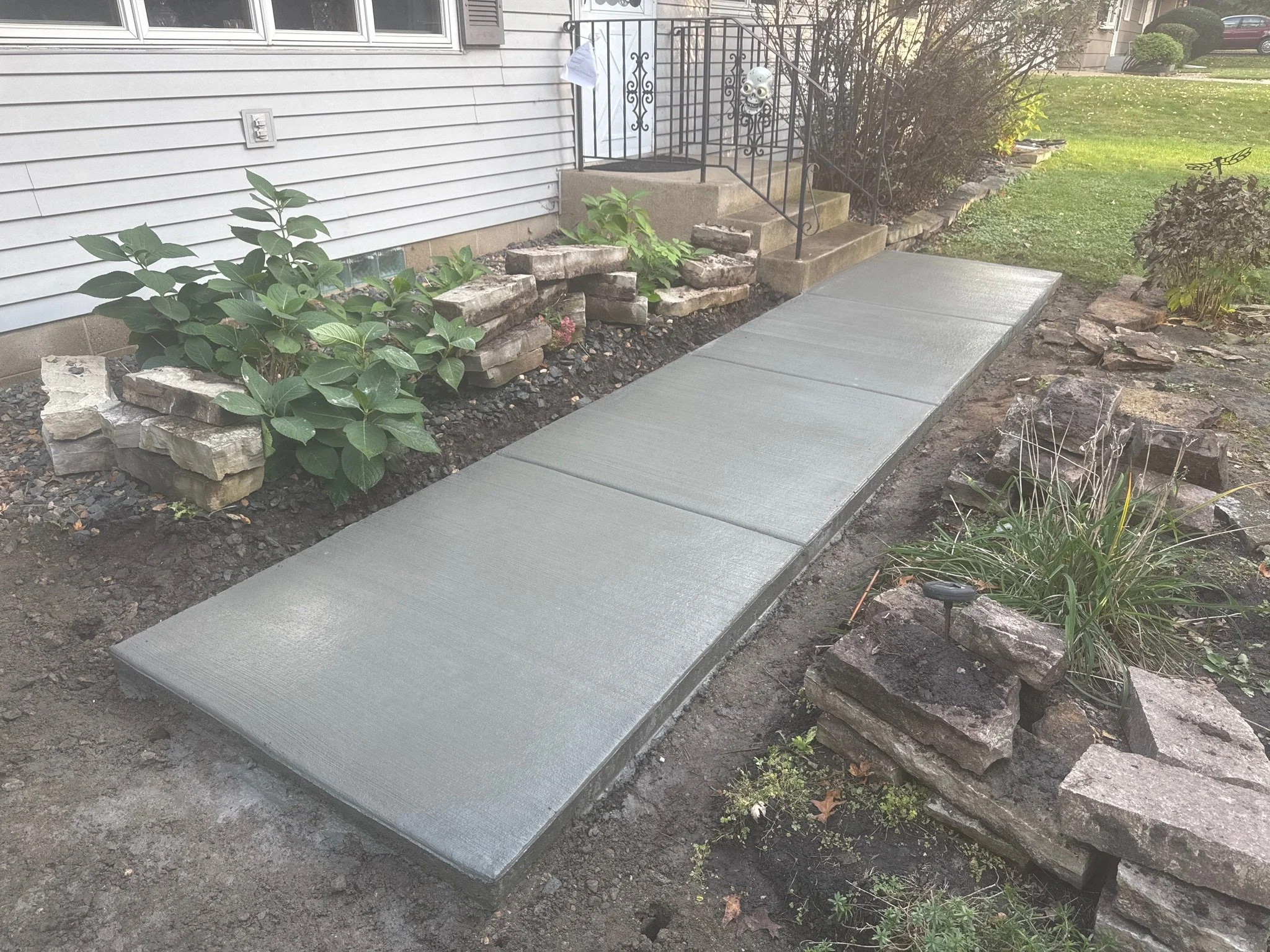 Freshly poured concrete walkway leading up to house front steps with a garden bed along the side containing green leafy plants and decorative rocks.