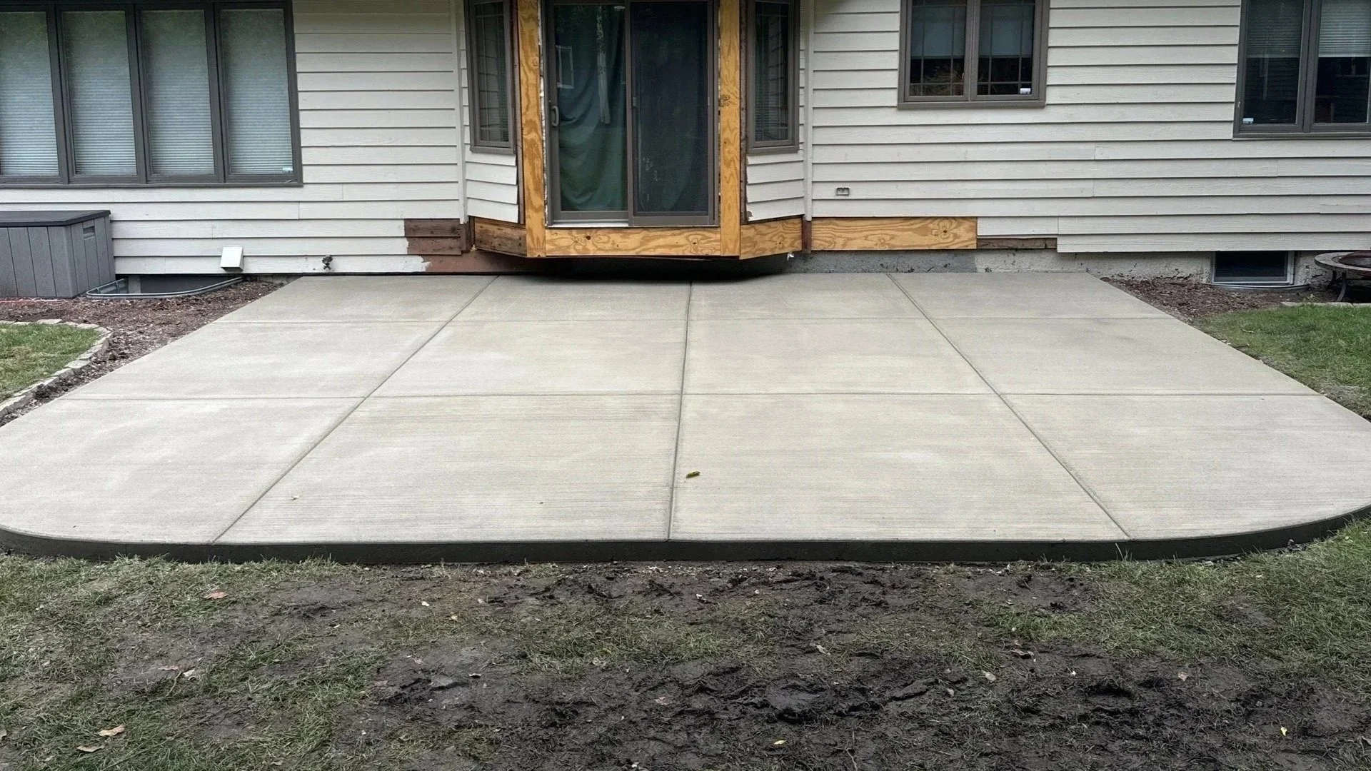 Newly poured concrete patio in front of house with siding, windows, and a glass sliding door, surrounded by grass and soil.