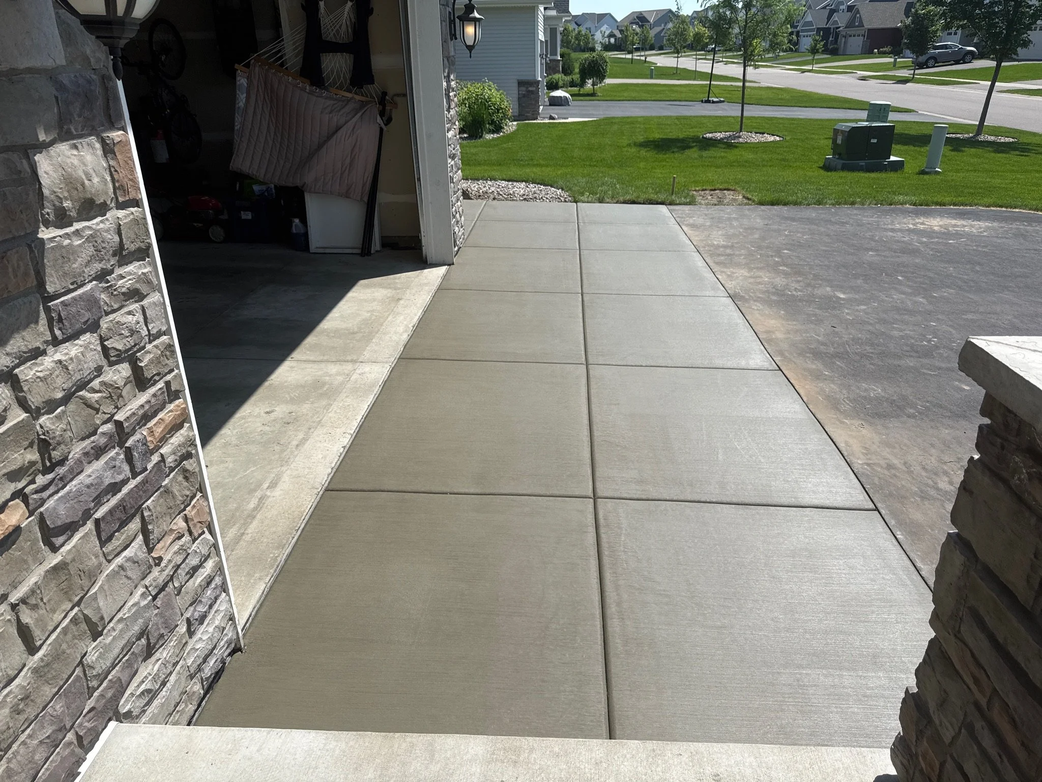 View of a newly poured concrete sidewalk in a residential driveway with a garage on the left, stone and brick wall on the right, and a well-manicured lawn with trees and houses in the background.