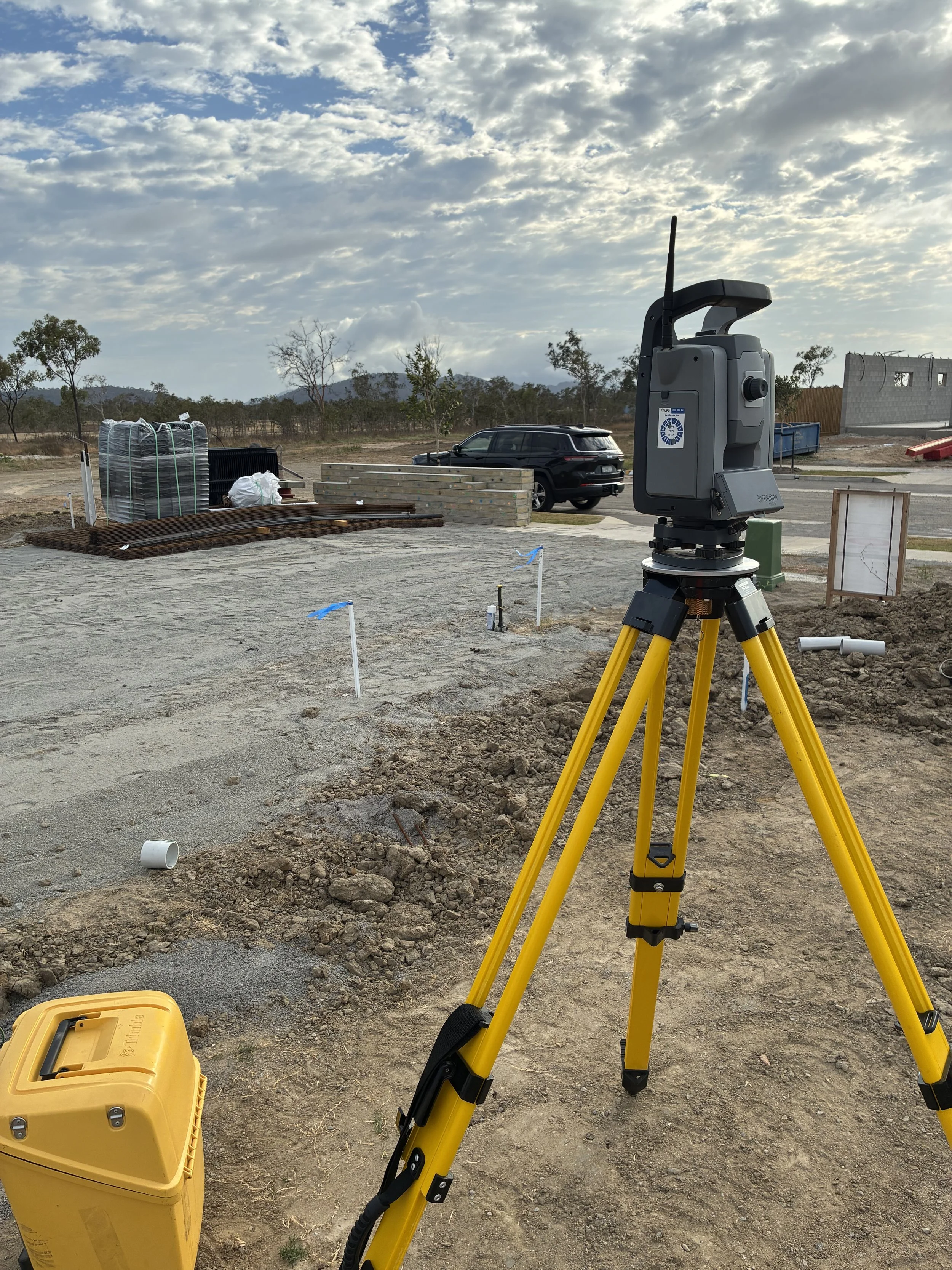 Land survey equipment, including a total station on a tripod, positioned at a construction site with dirt and construction materials in the background.