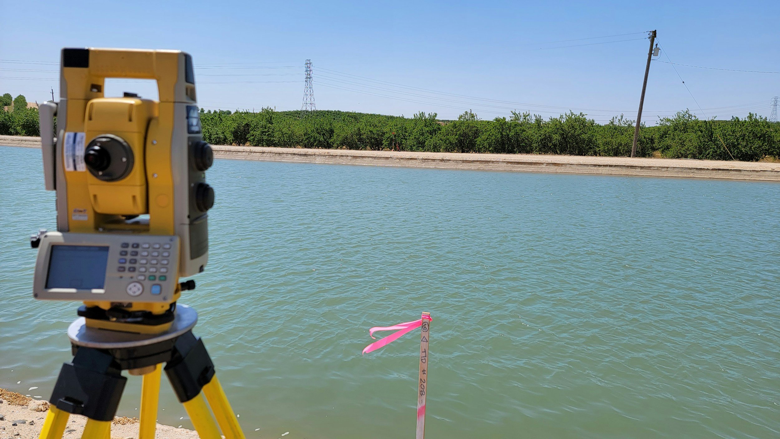 A surveying instrument set up on a tripod by a body of water, with a pink stakes marking measurement points on the water's edge, and a background of trees and power lines against a clear blue sky.