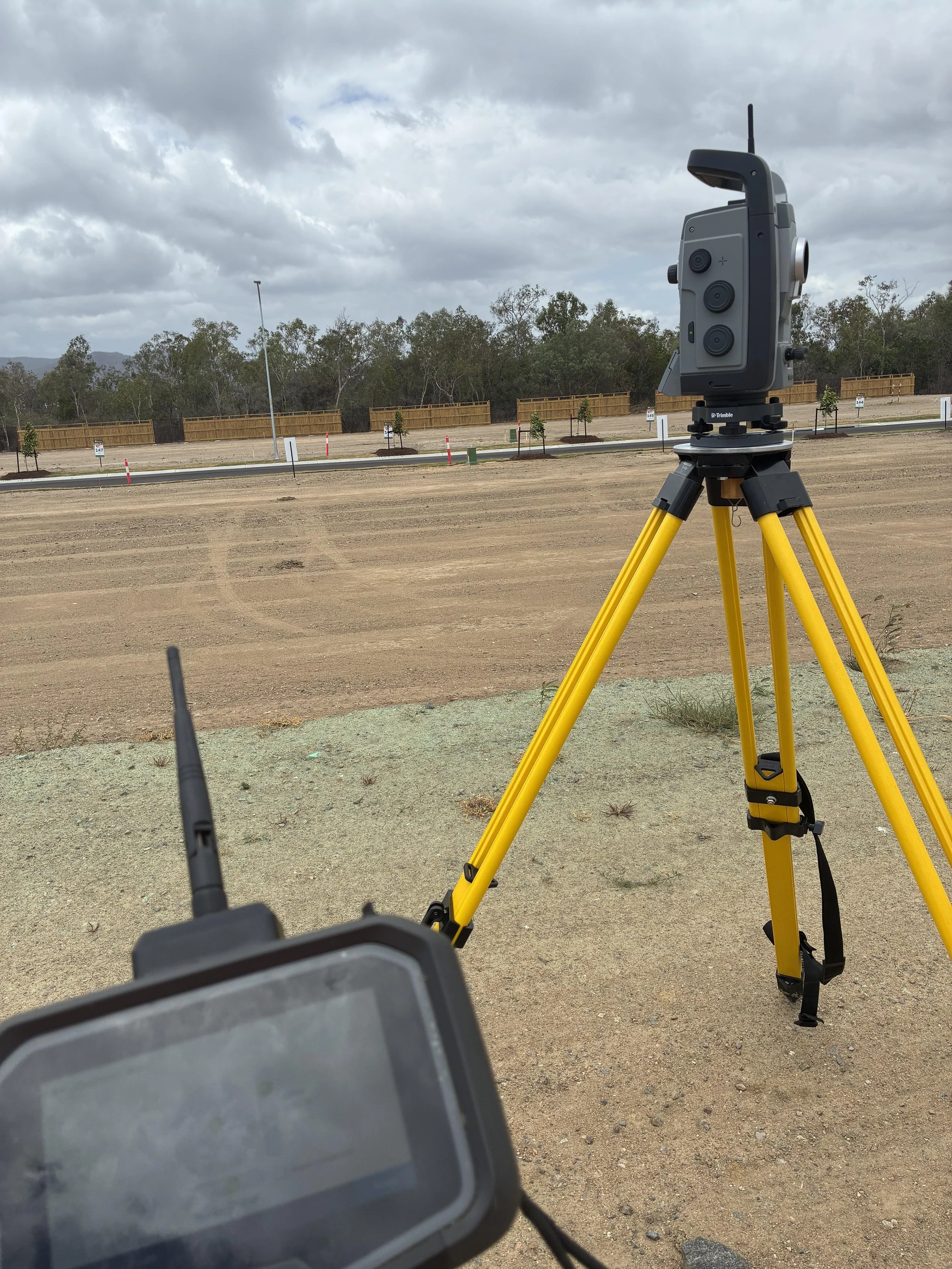 Construction site with a surveyor total station on a yellow tripod, a drone controller in the foreground, a tablet, and some small trees with mulch.