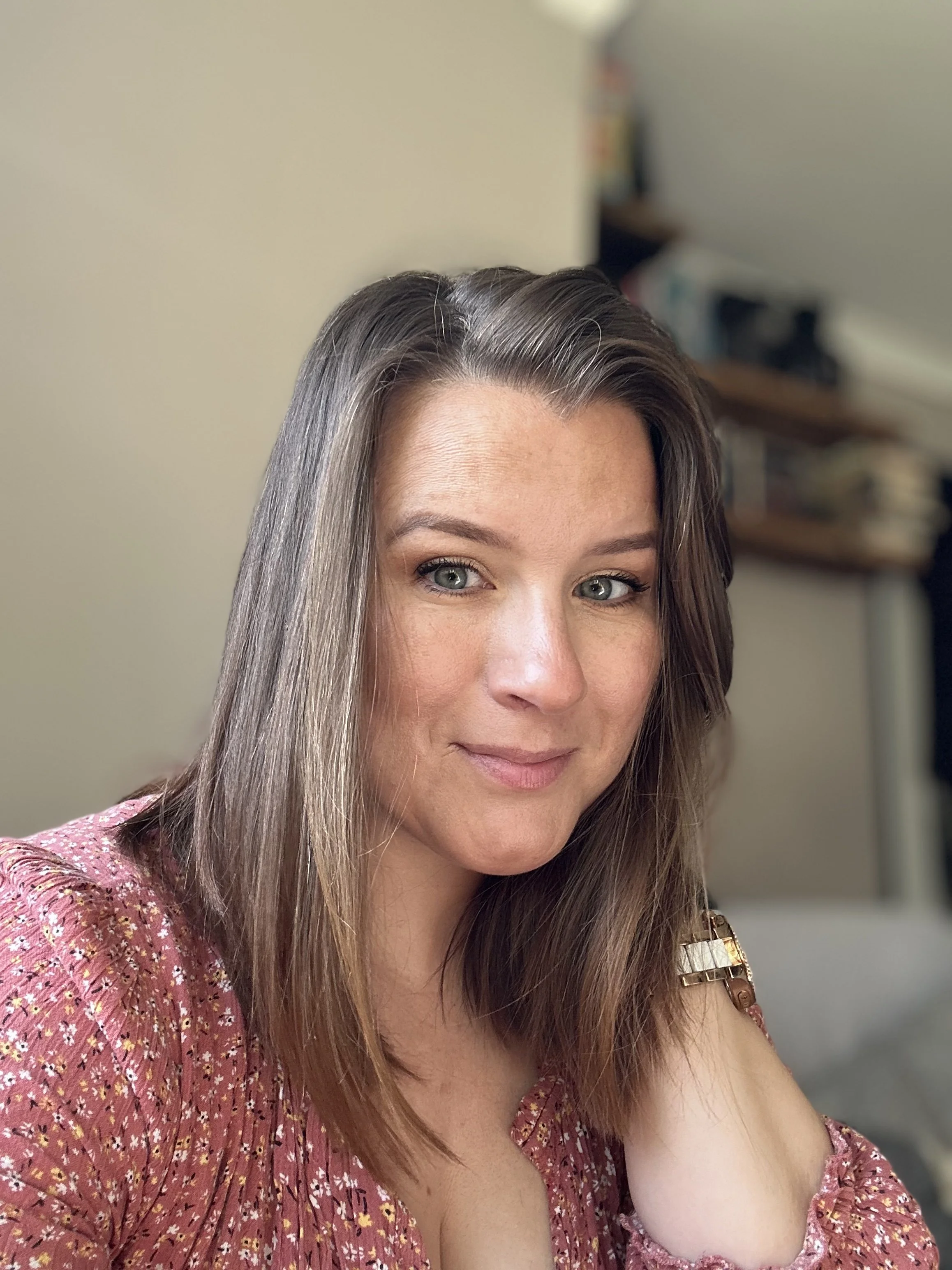 Close-up of a woman with shoulder-length brown hair, wearing a pink floral blouse, smiling in a room with beige walls and blurred background.