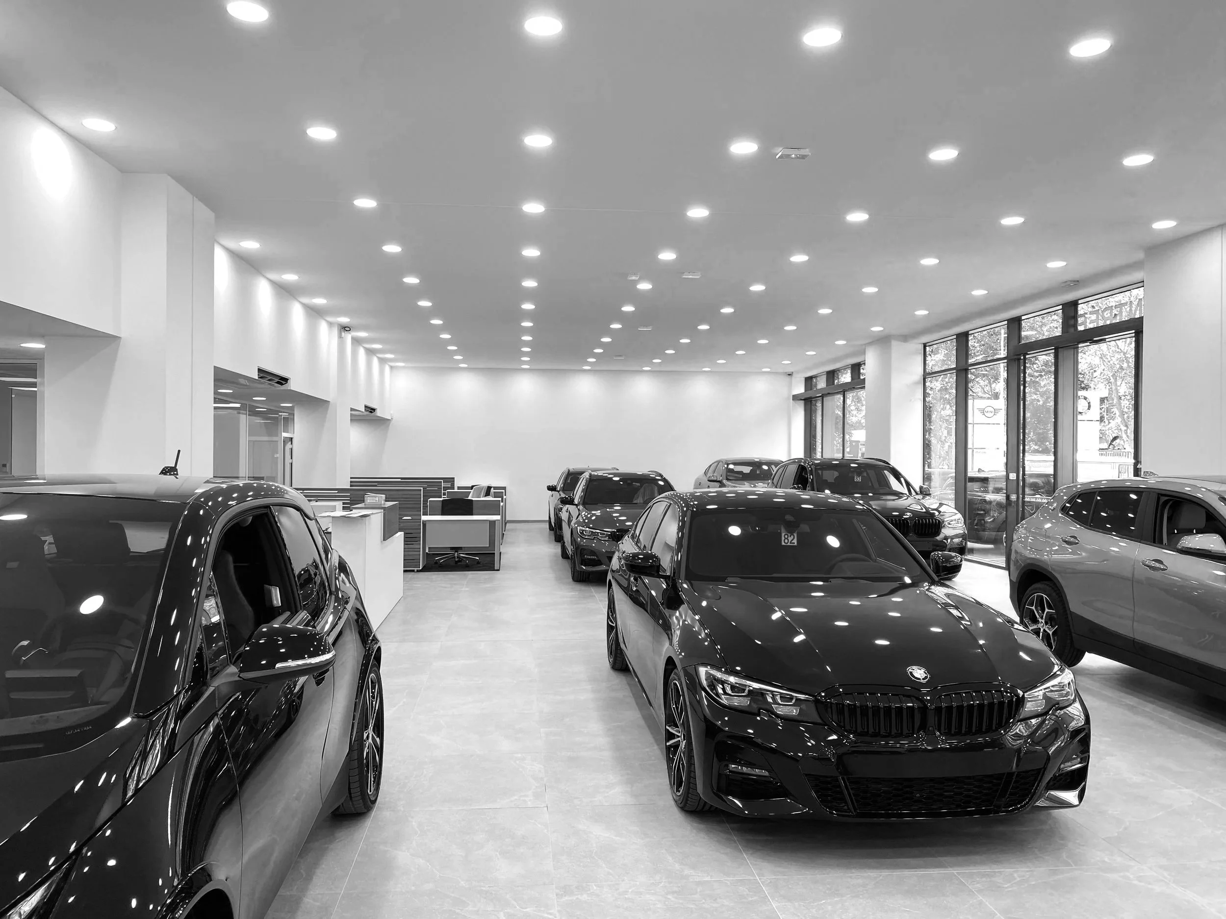 Intérieur d'un concessionnaire automobile avec plusieurs voitures modernes, dont une BMW noire au premier plan, et un espace de réception avec des bureaux en arrière-plan.