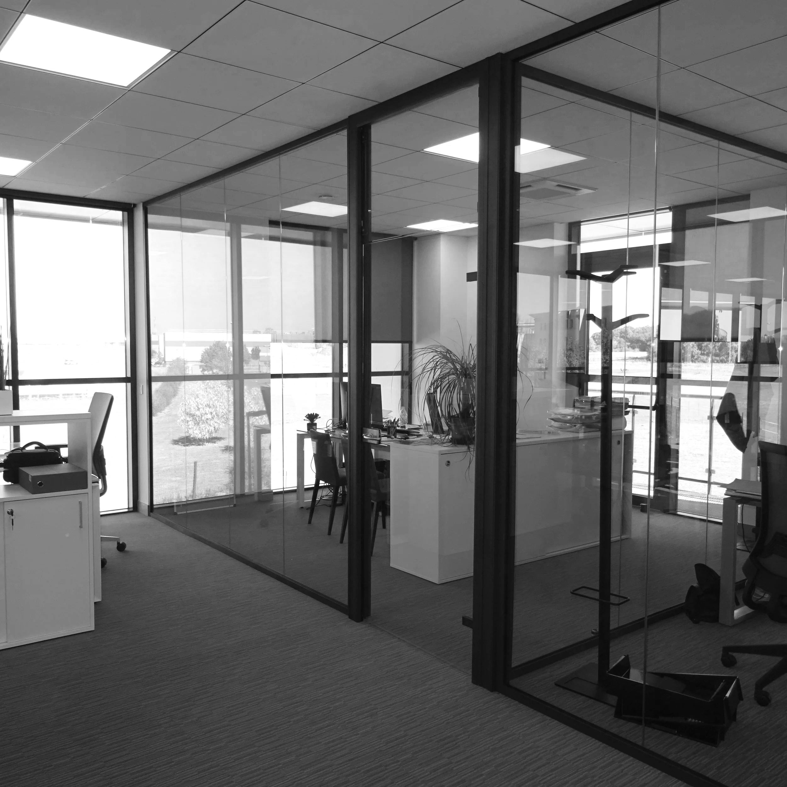 Bureau moderne avec murs en verre, mobilier de bureau et vue sur l’extérieur, éclairé par des lumières encastrées.