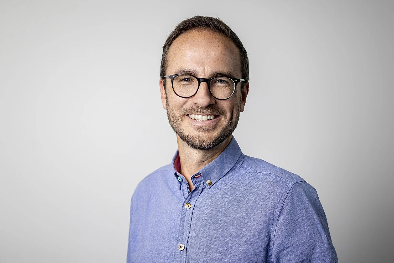 Porträt eines lächelnden Mannes mit Brille in einem blauen Hemd mit Knöpfen vor grauem Hintergrund.