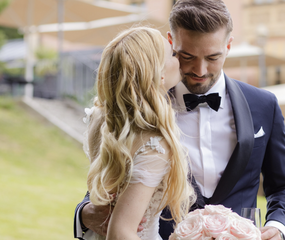 Ein Brautpaar bei einer Hochzeit, die Braut küsst den Bräutigam im Gesicht, der Bräutigam trägt einen schwarzen Anzug mit Fliege, die Braut hat lange blonde Haare und hält einen Blumenstrauß aus rosafarbenen Rosen.