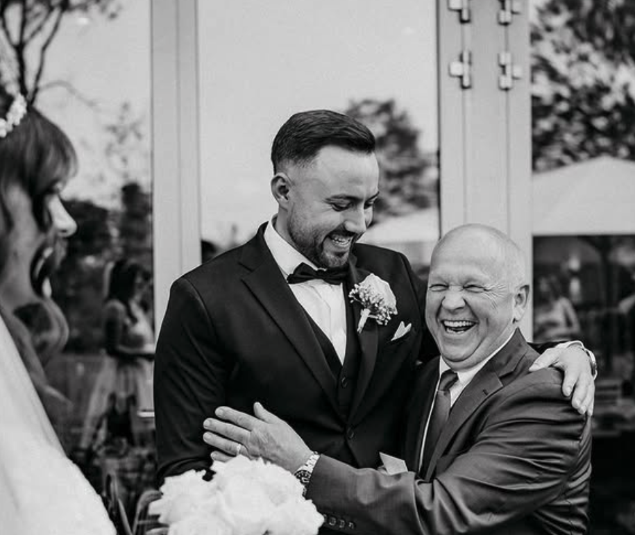 Zwei Männer bei einer Hochzeit, lachen und umarmen sich, im Hintergrund ein weiblicher Gast und eine geschlossene Tür.