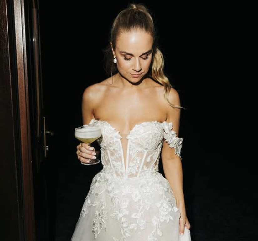 Braut in weißem Hochzeitskleid hält einen Cocktailglas, blickt nach unten, dunkler Hintergrund.
