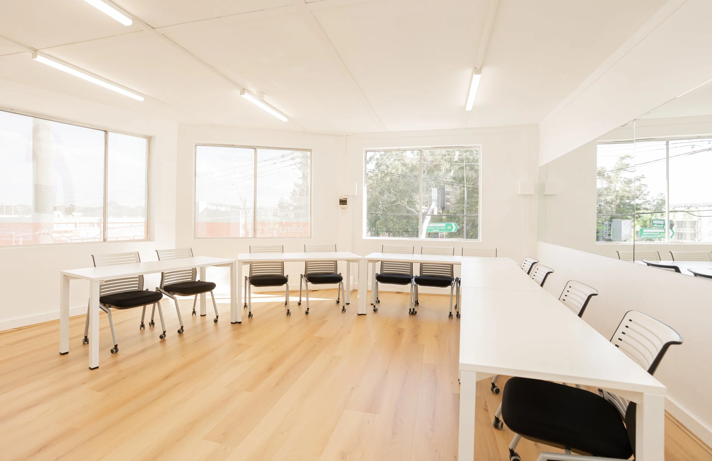 A bright conference room with white walls, wood floor, large windows, and a long white table surrounded by black chairs.