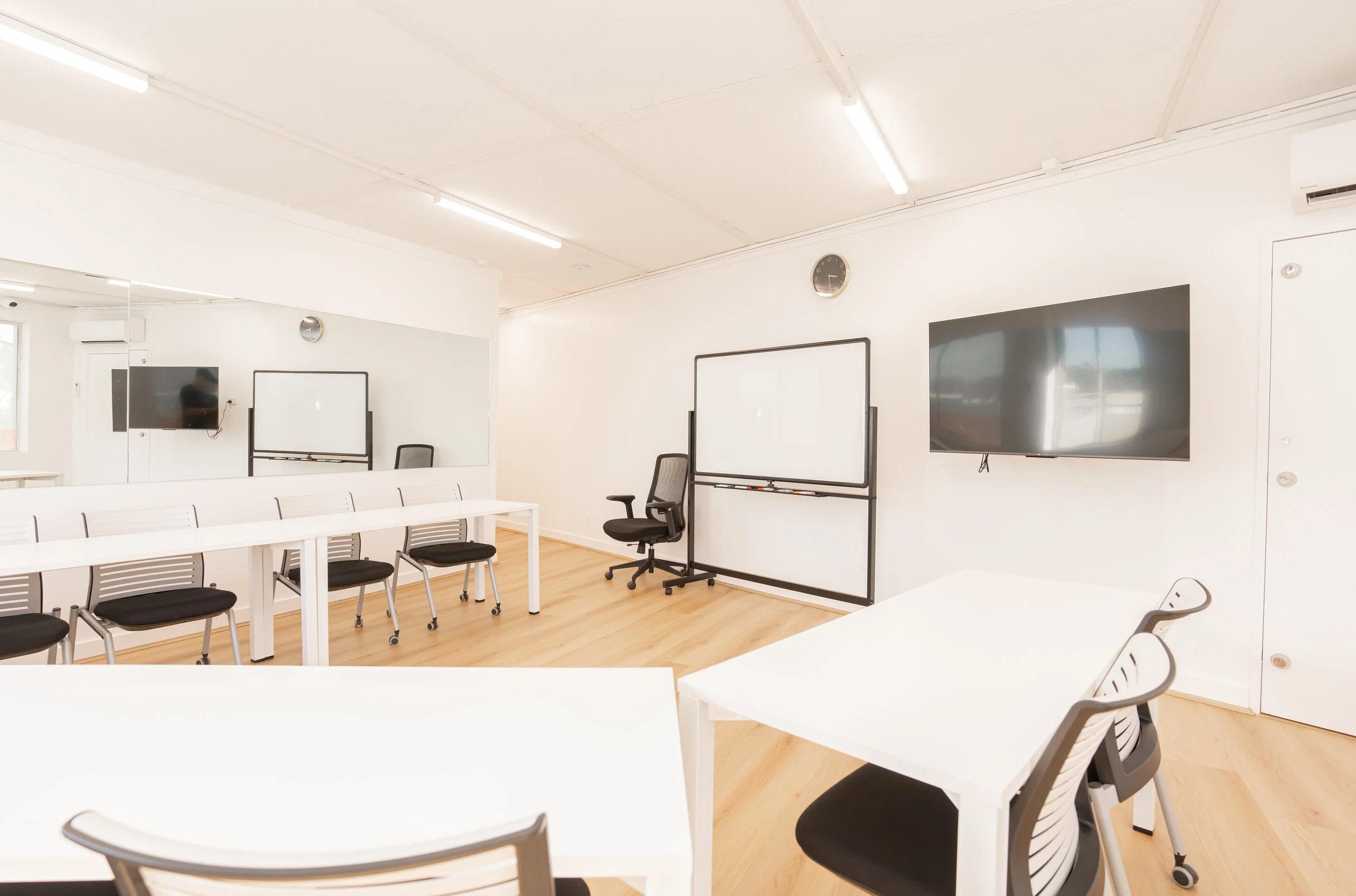 Empty classroom or meeting room with white walls, wooden floor, black chairs, white tables, a large mirror, a whiteboard, two wall-mounted screens, a clock, and an air conditioning unit.