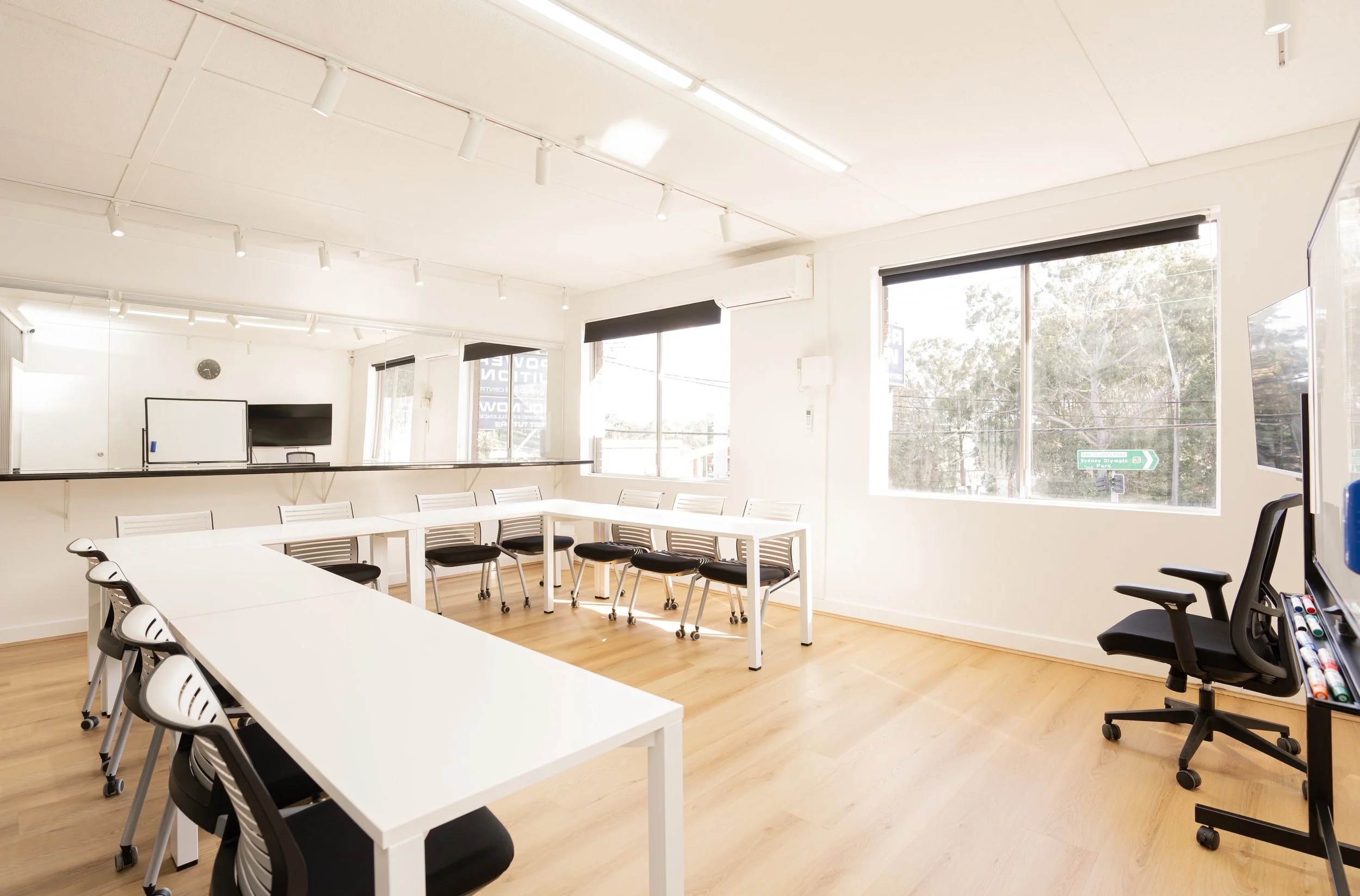 Empty modern conference or training room with white tables, black and white chairs, large windows, and a whiteboard with a TV mounted above it.