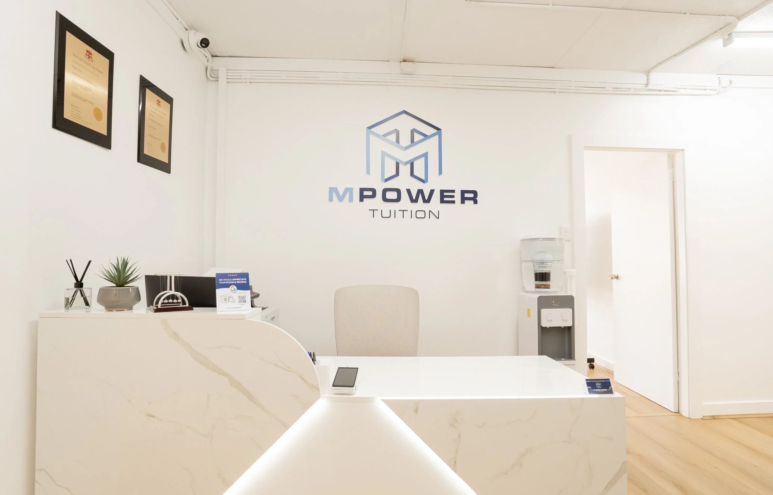 Modern reception area with white marble desk, a beige chair, water dispenser, and potted plant against a white wall with MPower Tuition logo and framed certificates.