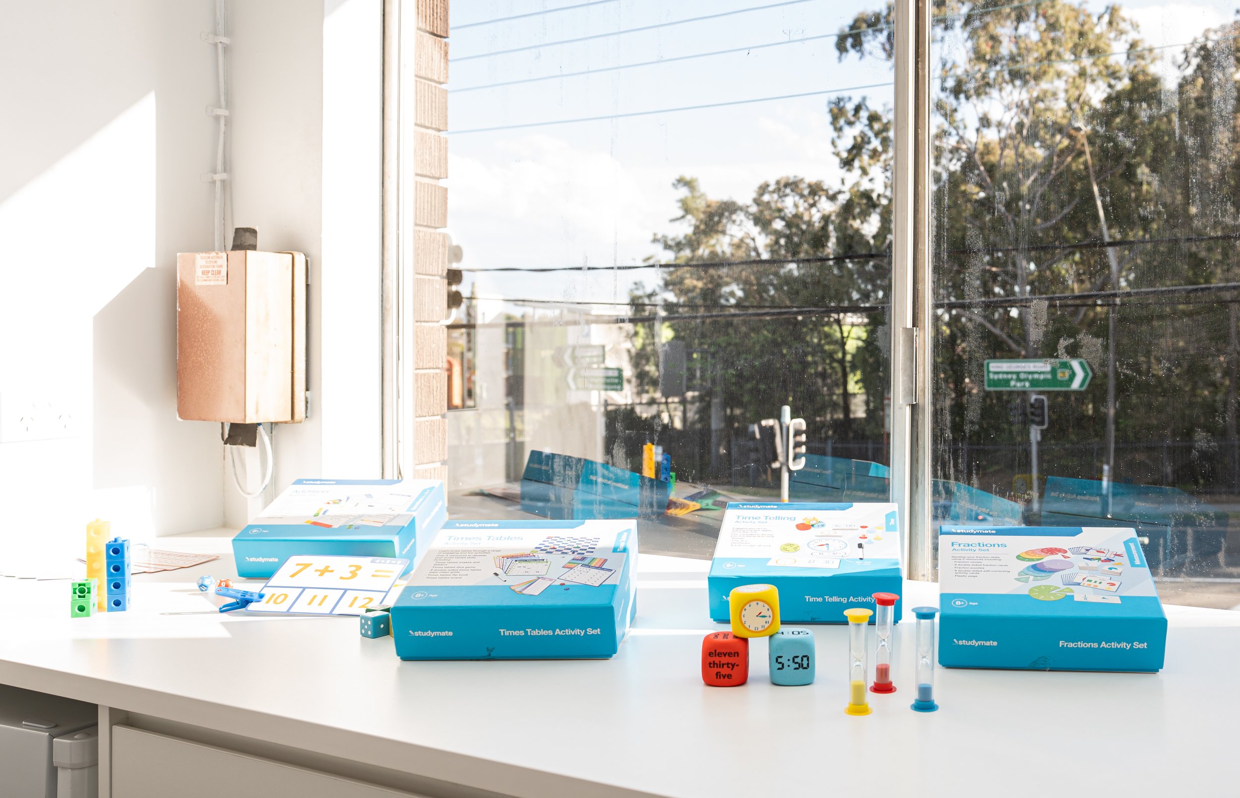 Educational toys and activities on a white table by a window, including teaching clocks, fraction activity sets, time telling activity set, dice, and hourglasses, with a sunny outdoor view.