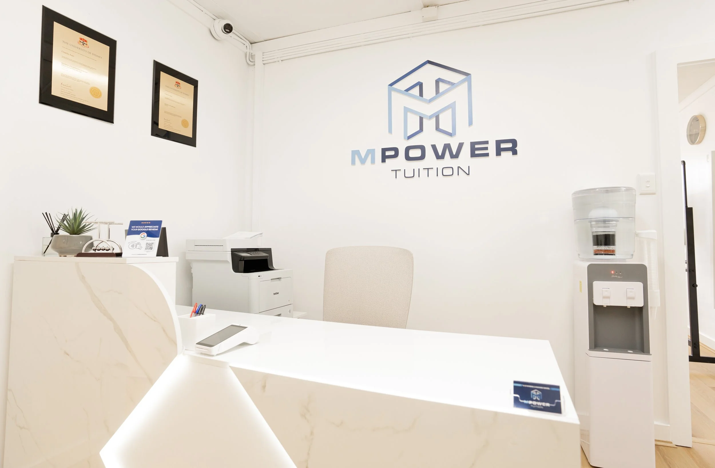Modern reception area with white marble desk, water cooler, framed certificates on the wall, and a logo that reads 'M POWER TUITION' on the wall.